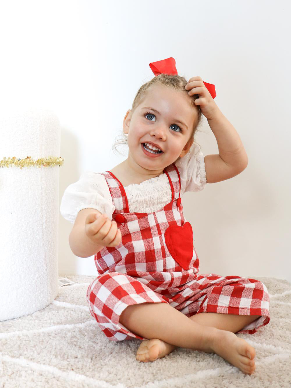 Child wearing a red and white checkered outfit with a bow, sitting on a white surface.