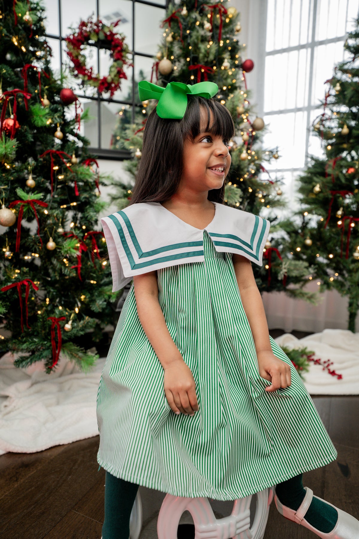 Young girl in a green and white dress with a bow in her hair, standing in front of Christmas trees.