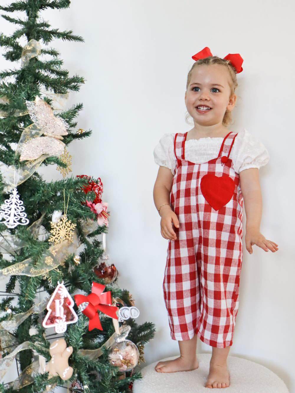 Child in a red checkered outfit with a heart design standing next to a decorated Christmas tree.