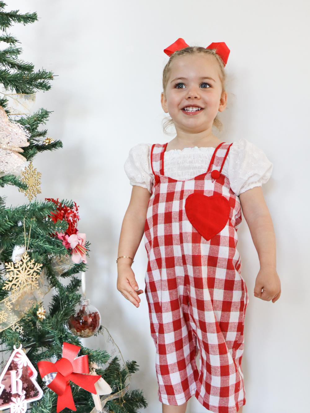 Child wearing a red and white checkered dress with a heart design, standing next to a decorated Christmas tree.