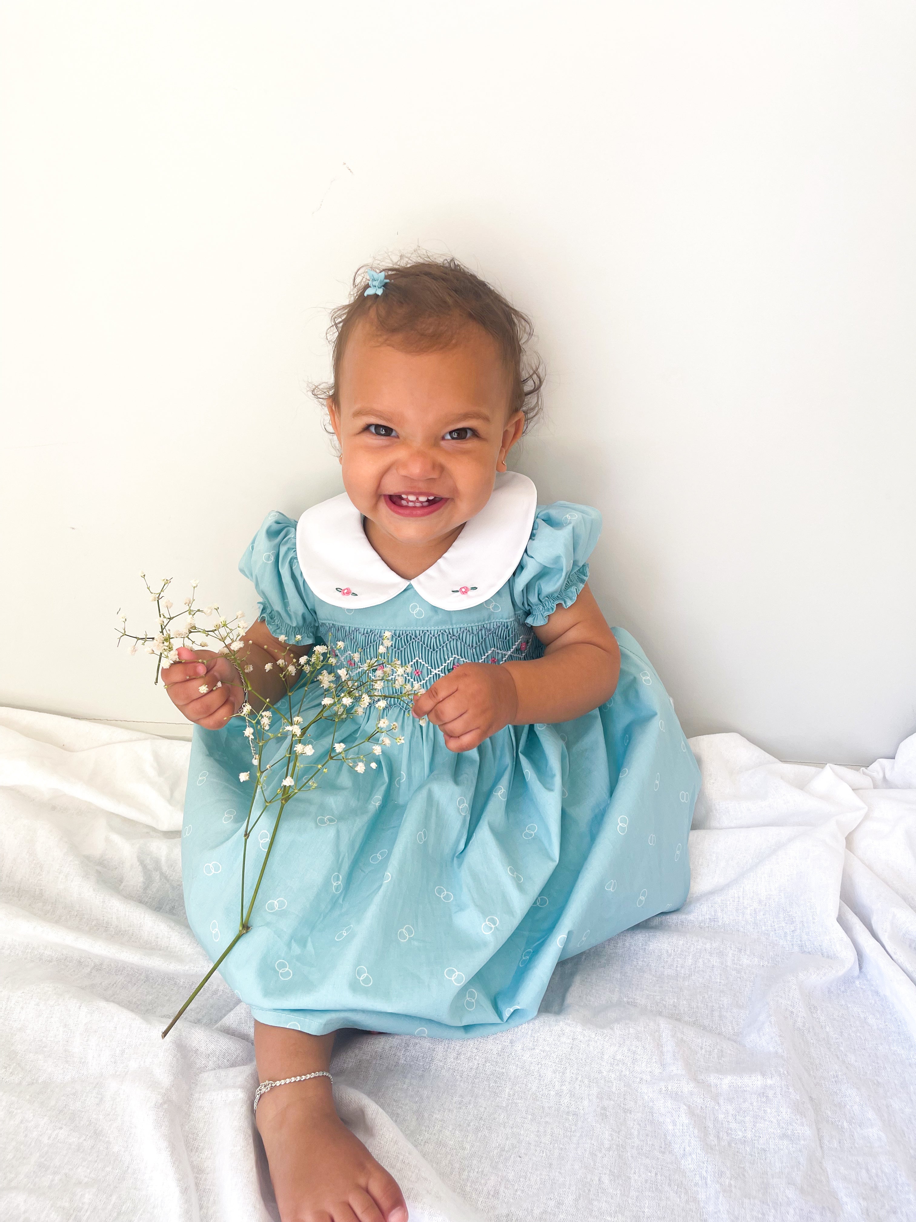 Baby wearing the Hand-smocked buebell Dress . Its a light blue dress with blue and white hand smocking with rose bud embroidery