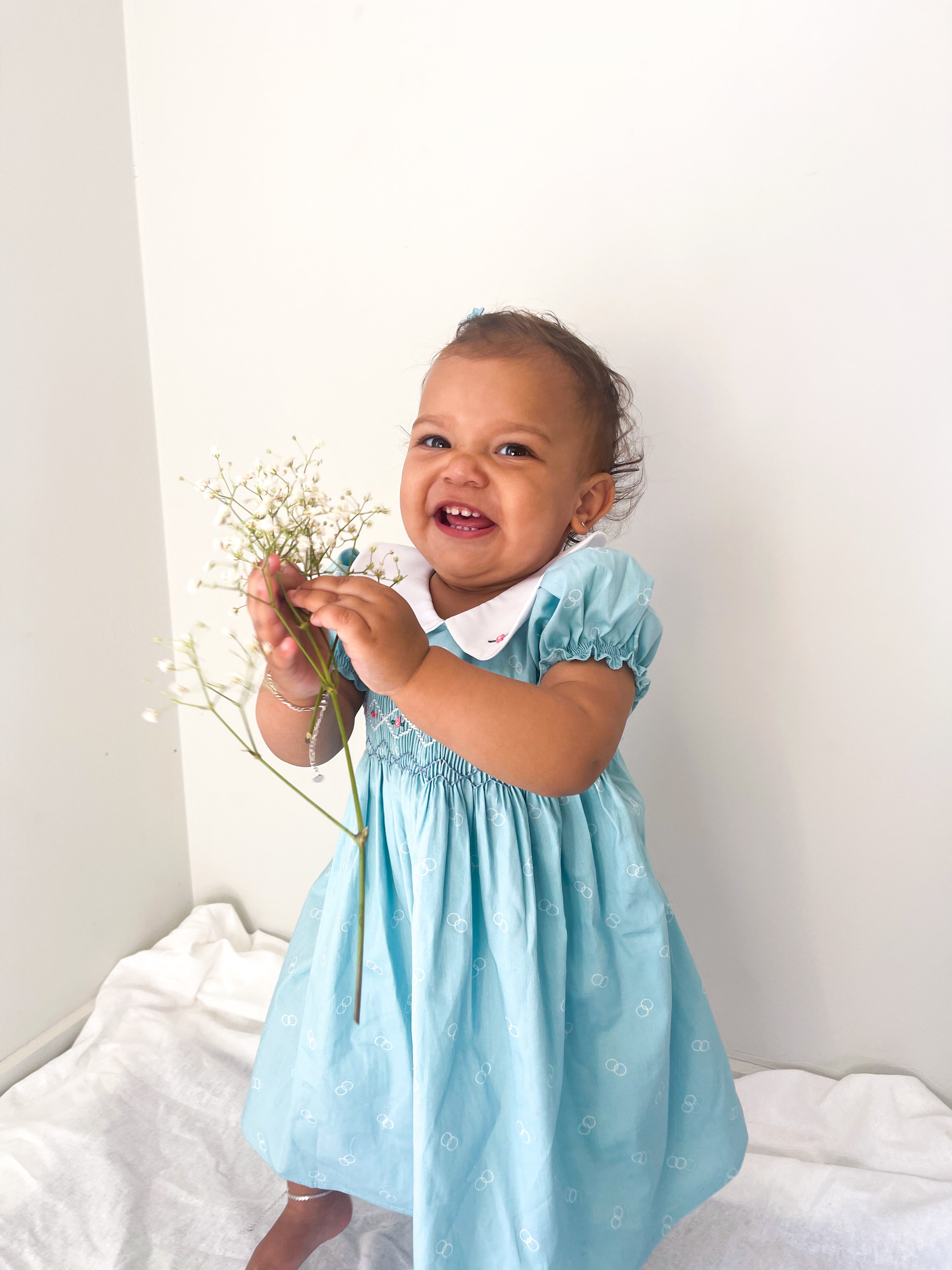 Baby wearing the light Blue and-smocked Bluebell Dress. Blue and white handsmocking with pink rose bud embrodiery.