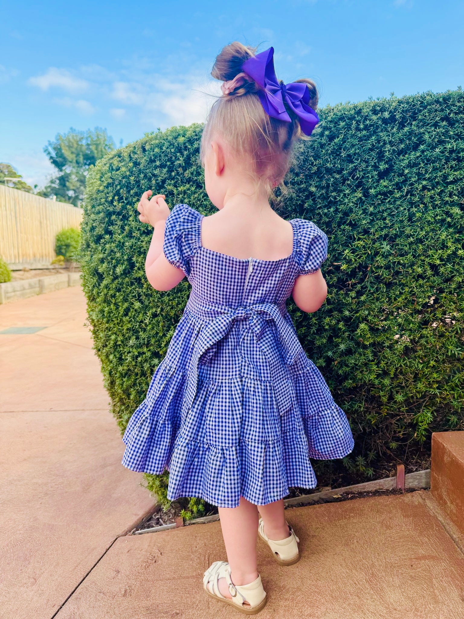 Back of the dress -Girl wearing purple and white gingham smock dress with pink bunnies in the middle, 3 tier ruffle dress. Back has a zip and bow stash