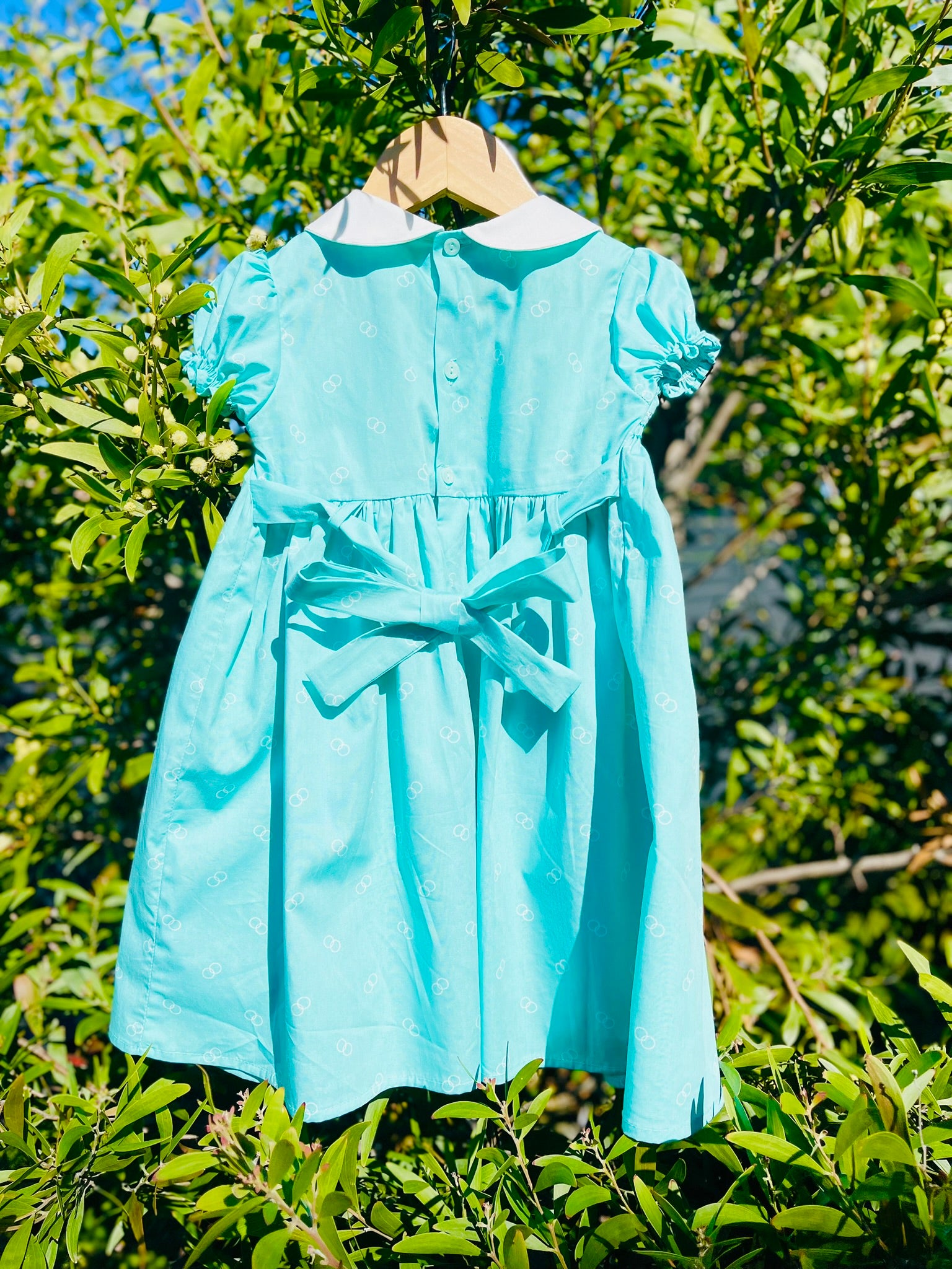 The Hand-smocked Emerald Rose Dress back which shows buttons and a bow stash. This is hanging on a green leafy tree. Emerald green and white handsmocking with pink rose bud embrodiery. White collar with one pink rose bud embrodiery on each collar.Dress is a green colour.