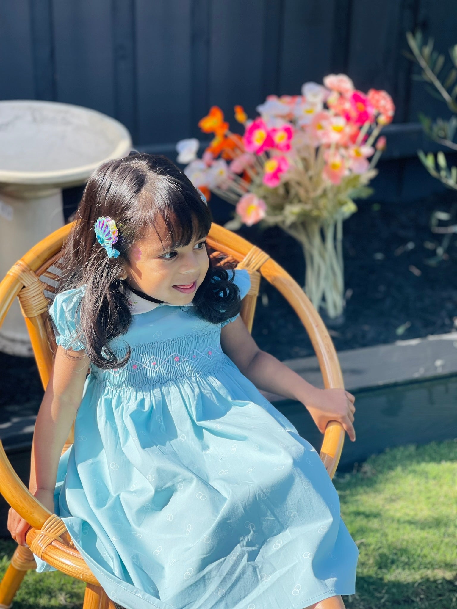 Girl sitting-Bluebell hand-smocked dress in blue with white collar and pink rose bud embroidery and blue white and pink handsmocking