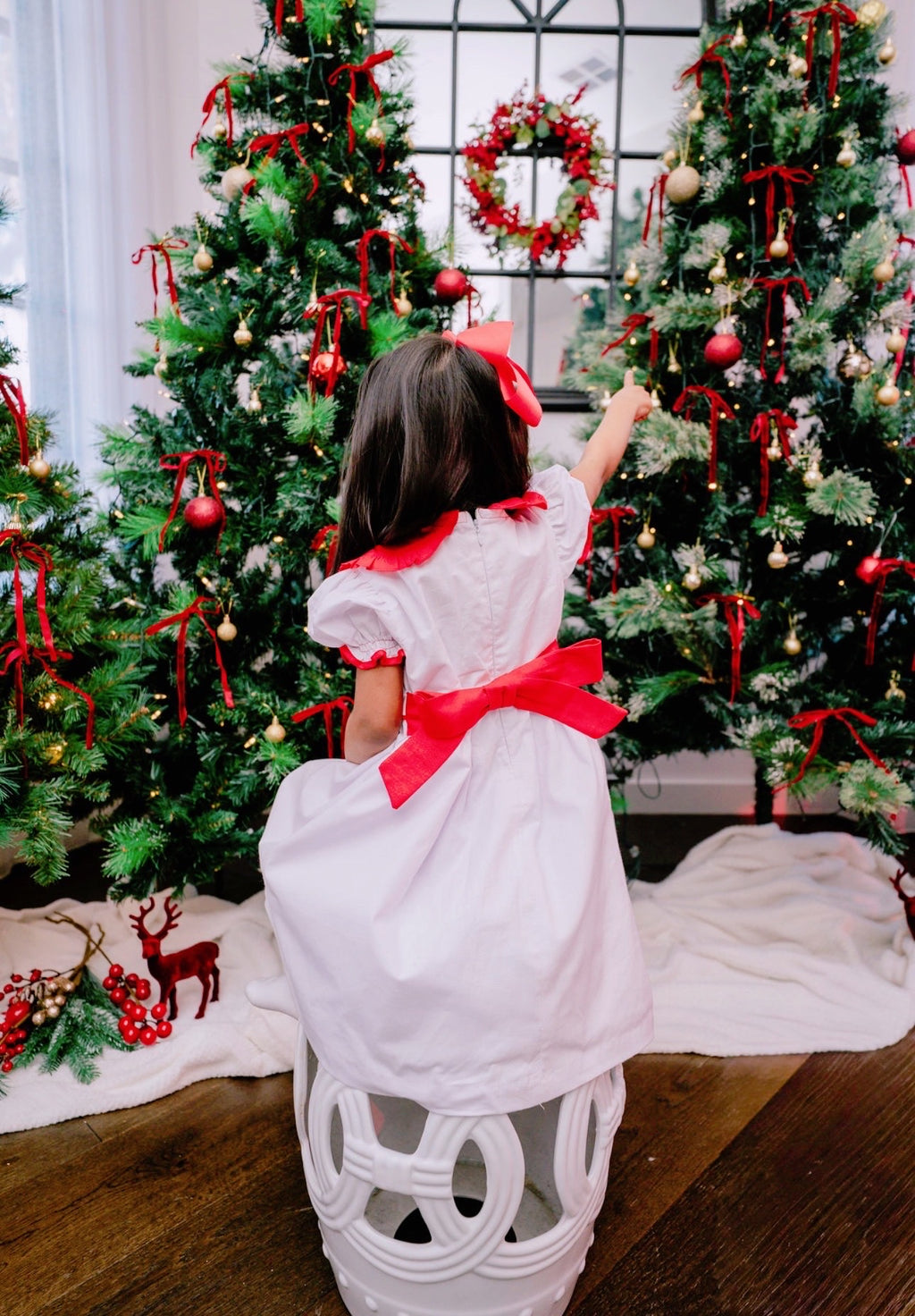 Child in a white dress with a red bow standing in front of Christmas trees.