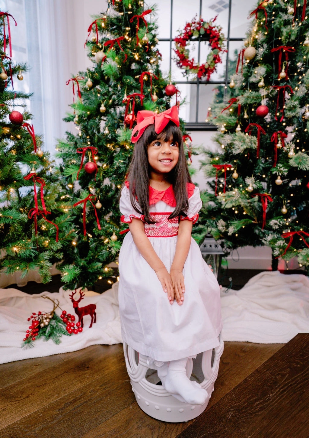 Young girl in a white dress with red accents sitting in front of Christmas trees.