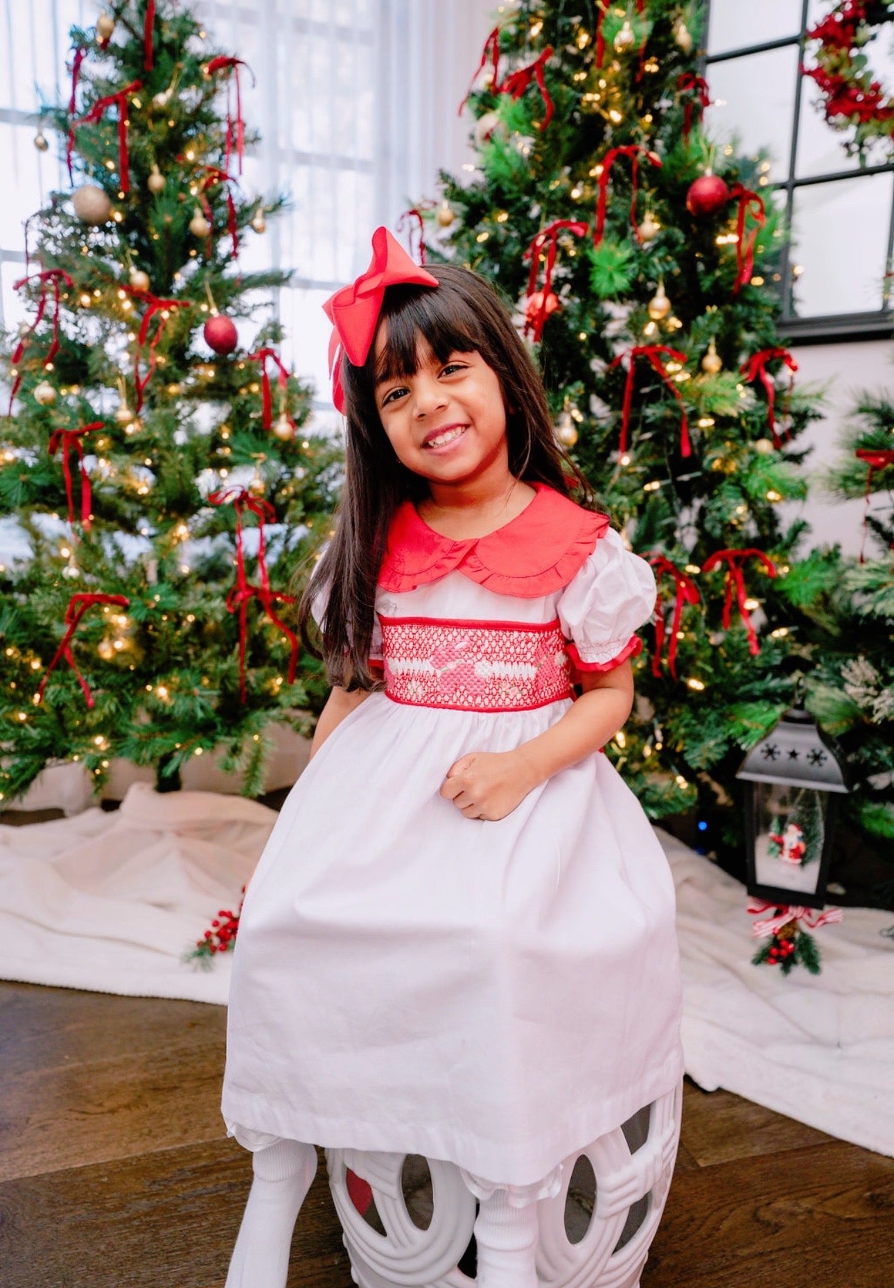 Young girl in a red and white dress standing in front of Christmas trees.