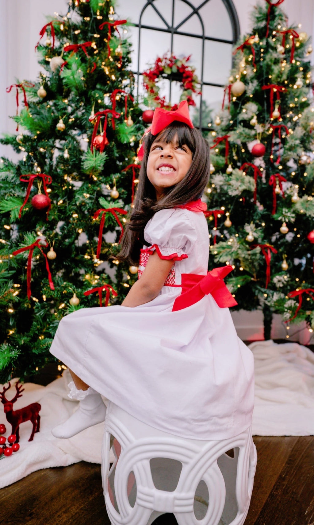 Child in a white dress with red bows sitting in front of Christmas trees.