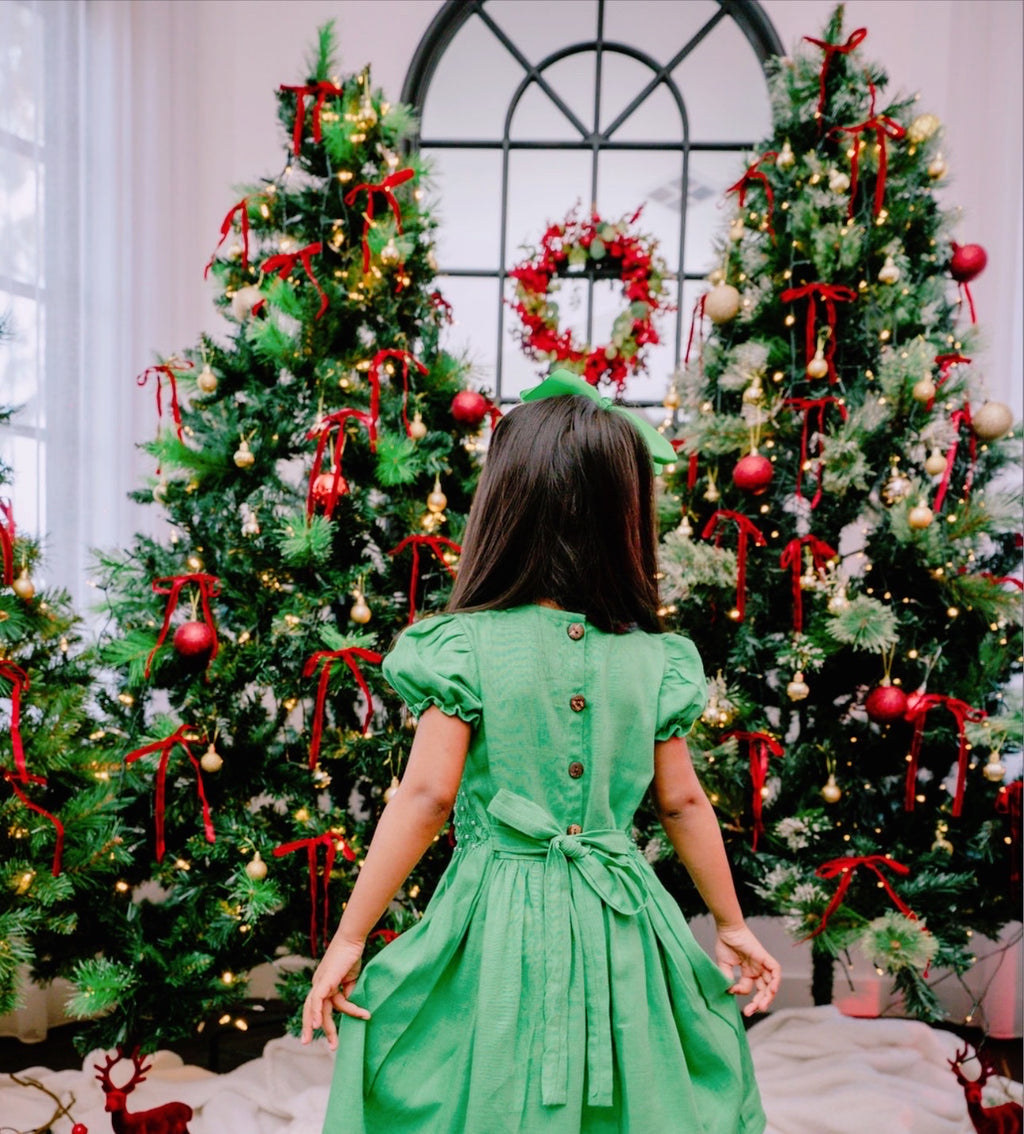 back shot of a toddler girl in front of Christmas trees wearing a green hand smocked dress