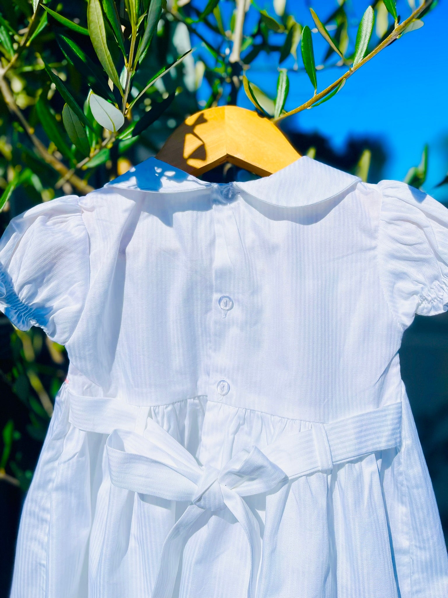 Close up and half of the back of the Hand-smocked Pure Blossom Dress. It has clear buttons and bow stash. The Dress is while and it has white, pink and green hand-smocking with dark pink rose bud embroidery. White collar with one pink rose bud embroidery on each collar. Dress is white in colour.