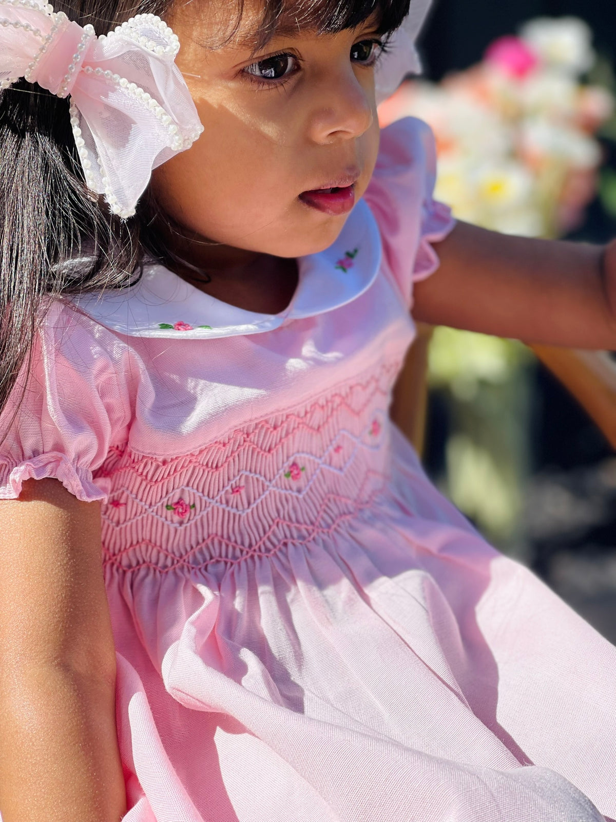 Close up shot of a girl while seated wearing the Hand-smocked Pink Sakura Dress. Light Pink Dress that has Pink and white handsmocking with dark pink rose bud embroidery. White collar with one pink rose bud embroidery on each collar. Dress is a light pink colour.