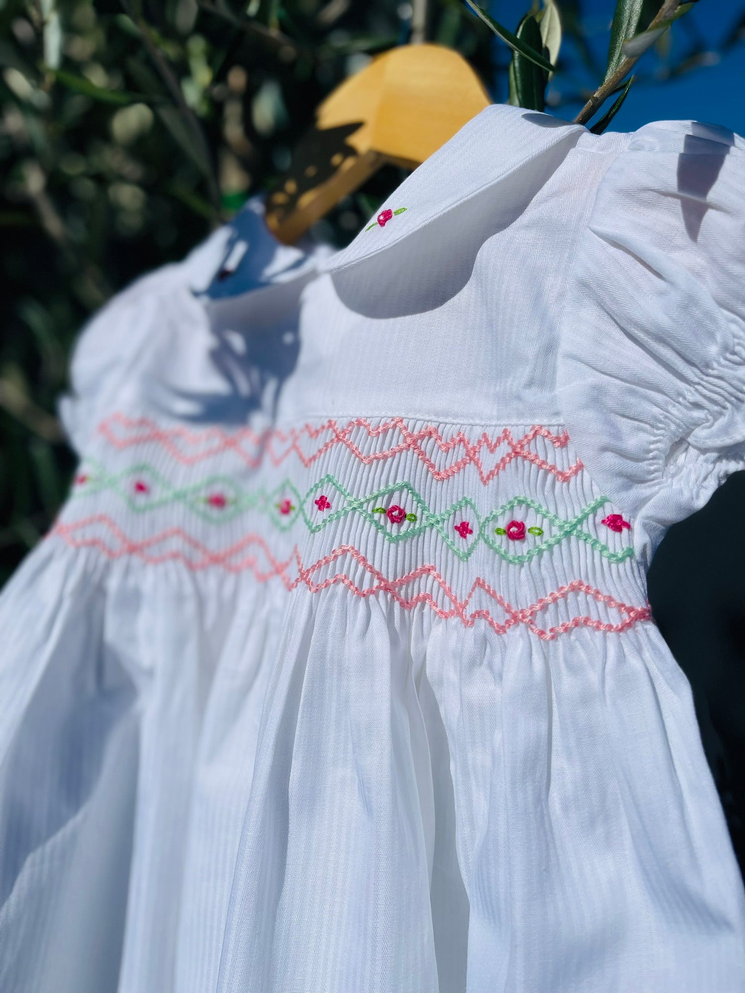 Close up photo of the top half of the Hand-smocked Pure Blossom Dress. The Dress is while and it has white, pink and green hand-smocking with dark pink rose bud embroidery. White collar with one pink rose bud embroidery on each collar. Dress is white in colour.