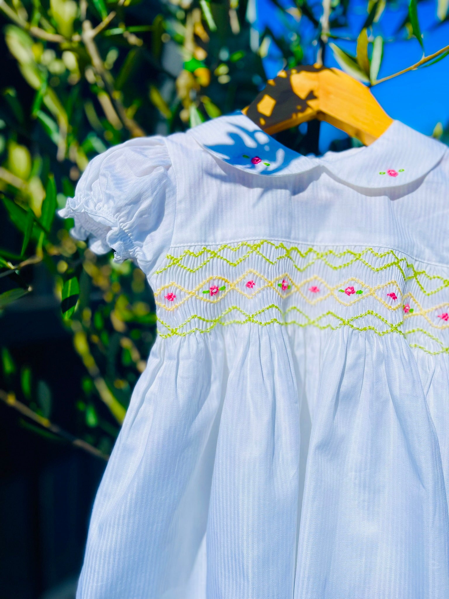 Front close up photo of the Hand-smocked Lily Blossom Dress. The Dress is while and it has white, green and yellow hand-smocking with dark pink rose bud embroidery. White collar with one pink rose bud embroidery on each collar. Dress is white in colour.