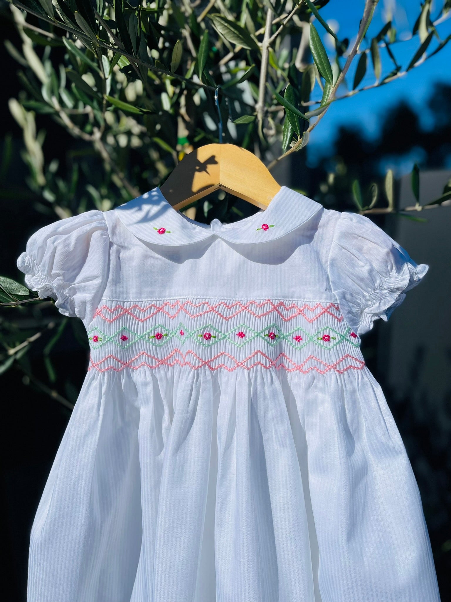 Front close up photo of the top half of the Hand-smocked Pure Blossom Dress. The Dress is while and it has white, pink and green hand-smocking with dark pink rose bud embroidery. White collar with one pink rose bud embroidery on each collar. Dress is white in colour.