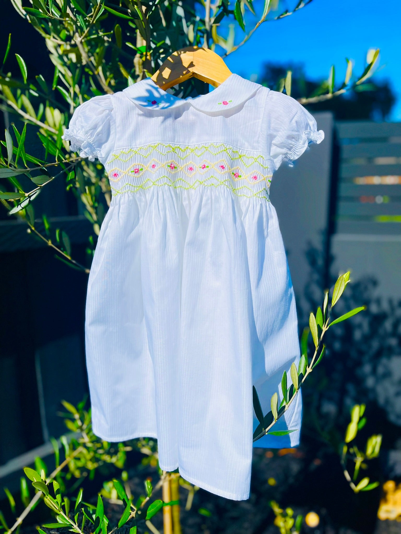 Front full length photo of The Hand-smocked Lily Blossom Dress. The Dress is while and it has white, green and yellow hand-smocking with dark pink rose bud embroidery. White collar with one pink rose bud embroidery on each collar. Dress is white in colour.