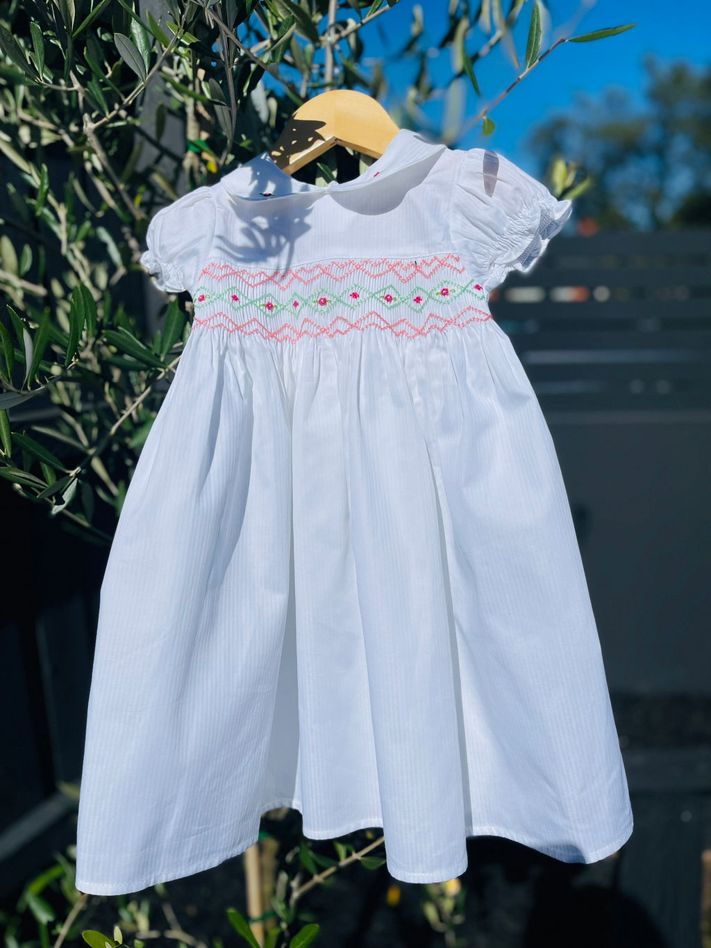 Front full length photo of the Hand-smocked Pure Blossom Dress. The Dress is while and it has white, pink and green hand-smocking with dark pink rose bud embroidery. White collar with one pink rose bud embroidery on each collar. Dress is white in colour.