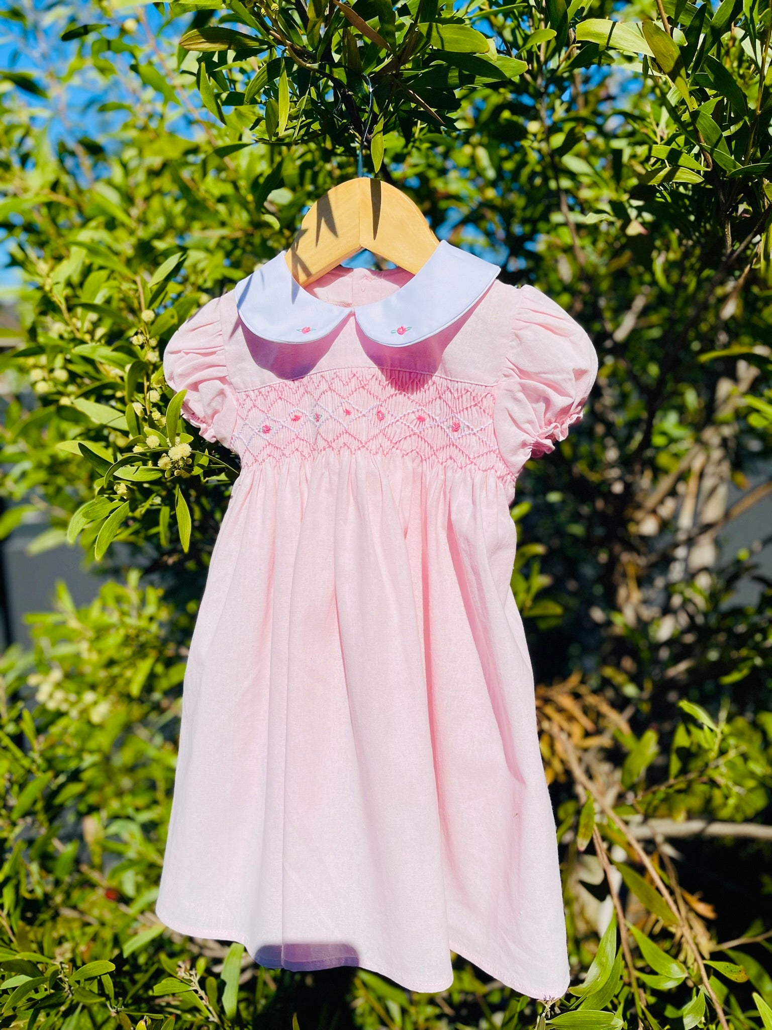 Front of the hand-smocked Pink Sakura Dress. Light Pink Dress that has Pink and white handsmocking with dark pink rose bud embroidery. White collar with one pink rose bud embroidery on each collar. Dress is a light pink colour.