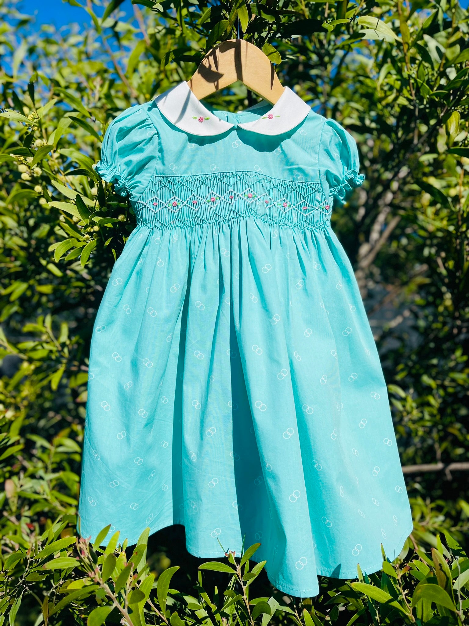 Photo of the Hand-smocked Emerald Rose hanging on a leafy green branch Dress. Emerald green and white handsmocking with pink rose bud embrodiery. White collar with one pink rose bud embrodiery on each collar.Dress is a green colour.
