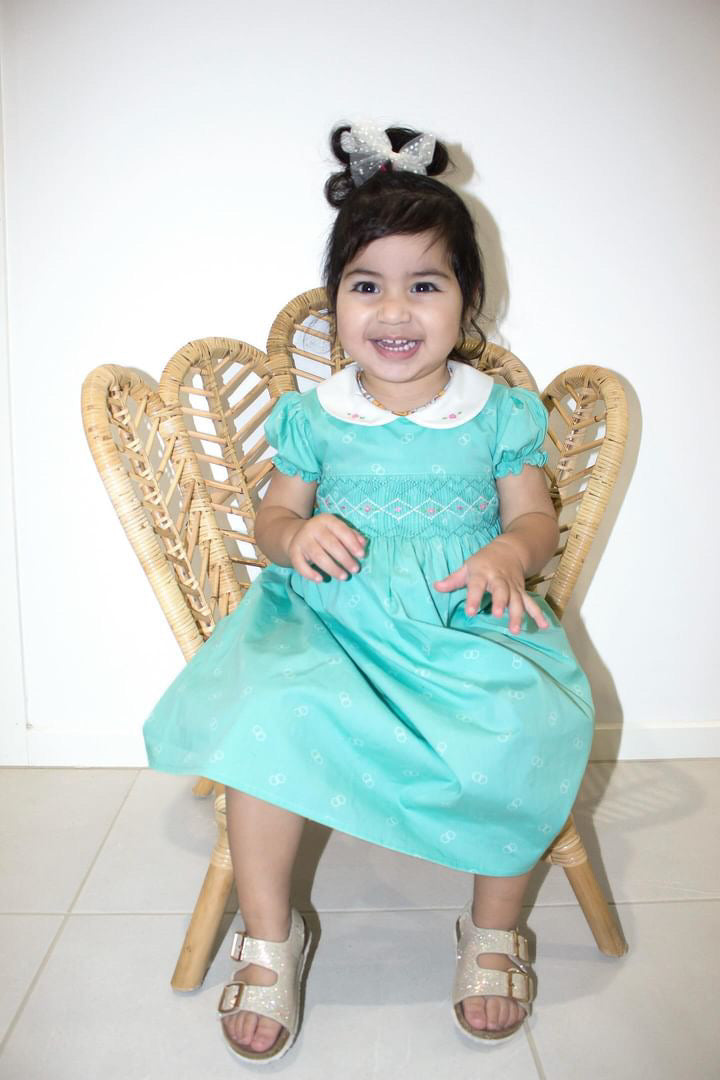 Girl Toddler wearing the Hand-smocked Emerald Rose Dress. Emerald green and white handsmocking with pink rose bud embrodiery. White collar with one pink rose bud embrodiery on each collar.Dress is a green colour.