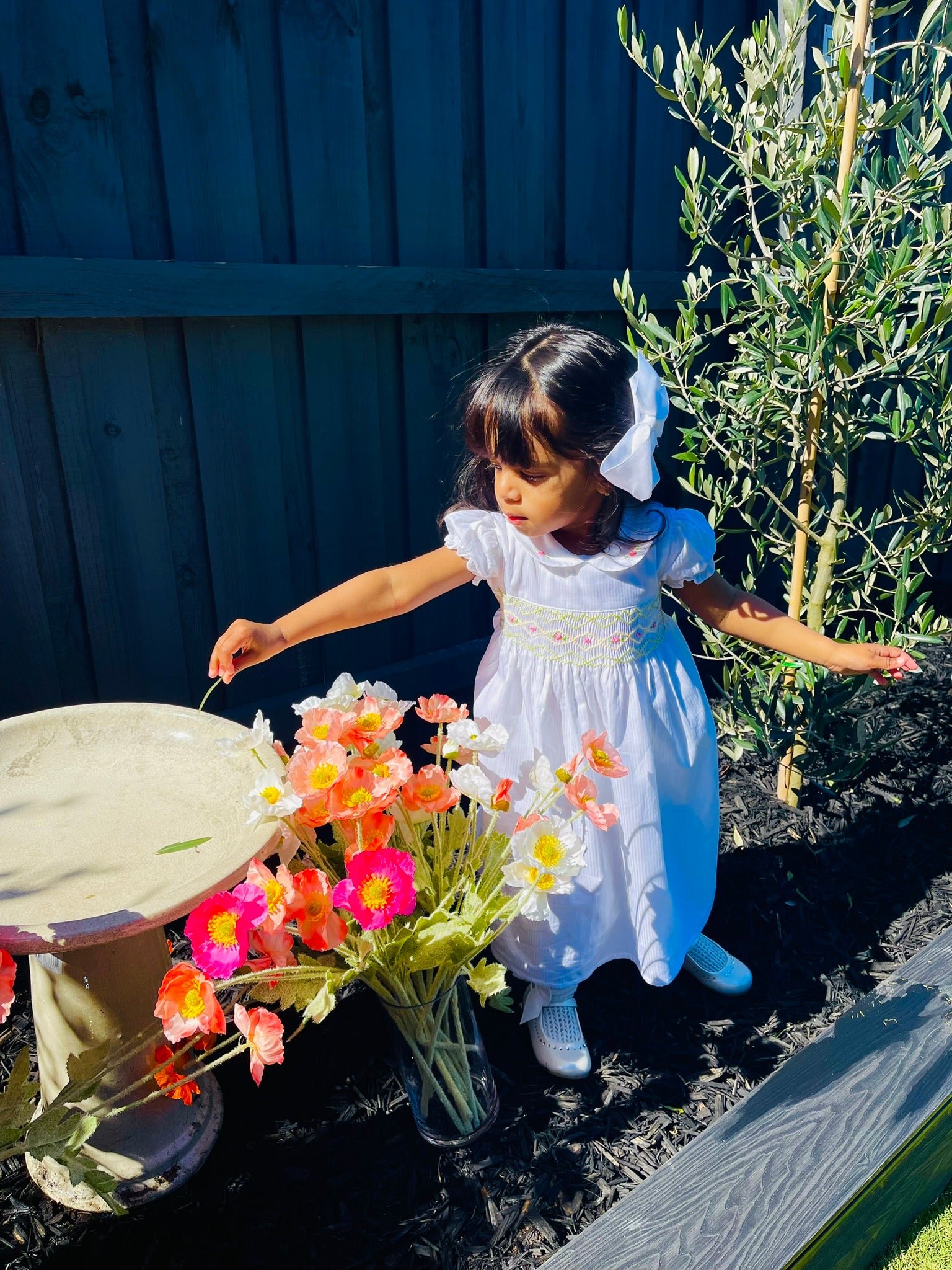 Girl Toddler wearing The Hand-smocked Lily Blossom Dress playing with flowers and leaves. The Dress is while and it has white, green and yellow hand-smocking with dark pink rose bud embroidery. White collar with one pink rose bud embroidery on each collar. Dress is white in colour.