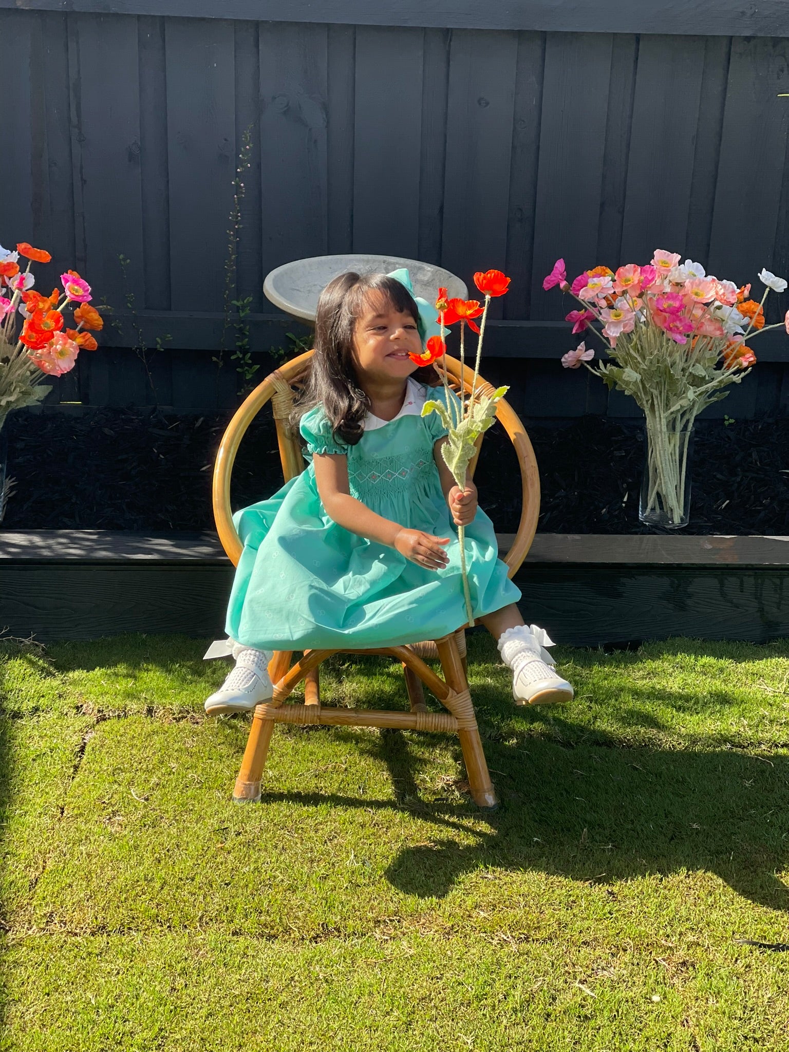 Girl Toddler blowing on orange flowers wearing the Hand-smocked Emerald Rose Dress. Emerald green and white handsmocking with pink rose bud embrodiery. White collar with one pink rose bud embrodiery on each collar.Dress is a green colour.