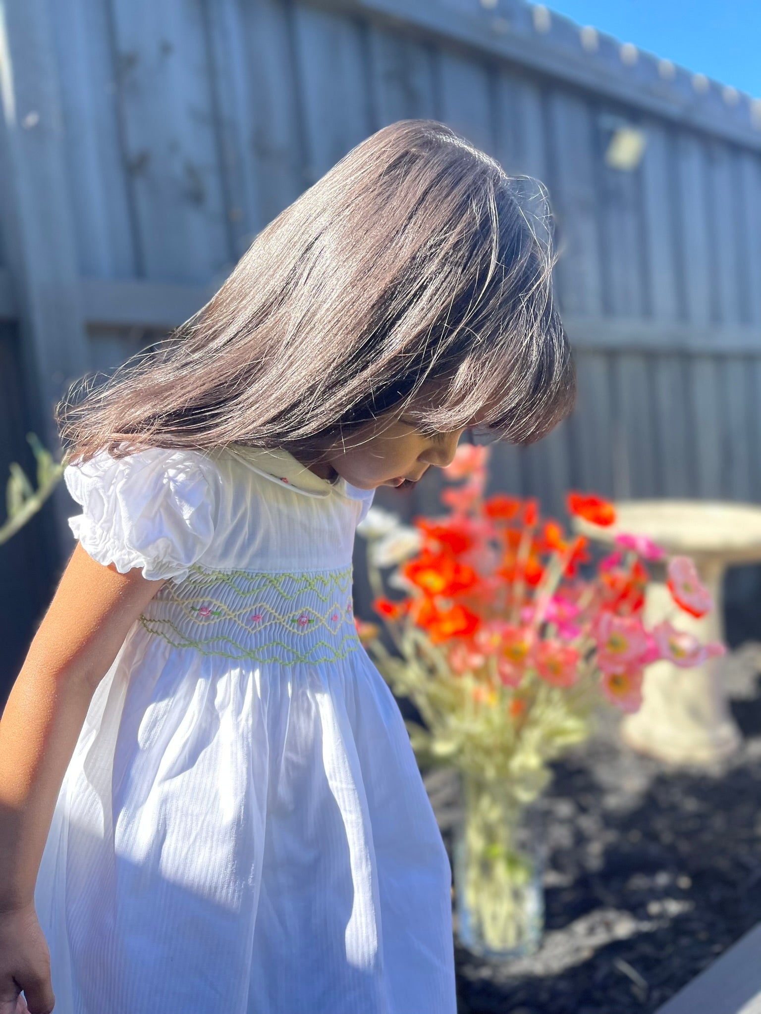 Girl looking down at her dress which is the hand-smocked Lily Blossom Dress. The Dress is while and it has white, green and yellow hand-smocking with dark pink rose bud embroidery. White collar with one pink rose bud embroidery on each collar. Dress is white incolour.