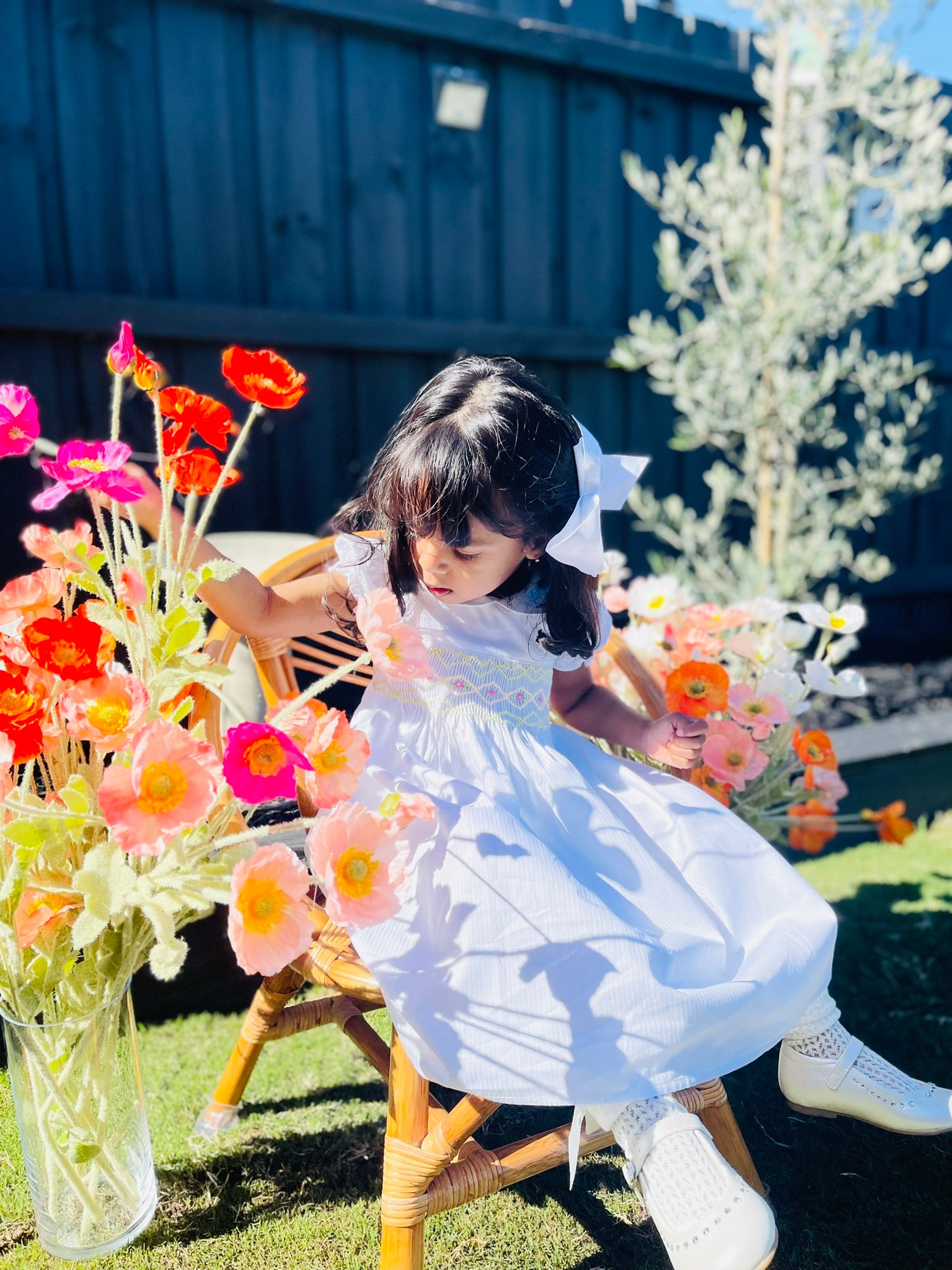 Girl sitting and playing with a flower vase wearing the Hand-smocked Lily Blossom Dress. The Dress is while and it has white, green and yellow hand-smocking with dark pink rose bud embroidery. White collar with one pink rose bud embroidery on each collar. Dress is white in colour.
