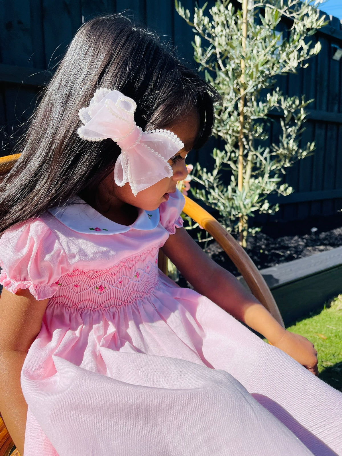 Girl toddler looking down while seated wearing the Hand-smocked Pink Sakura Dress. Light Pink Dress that has Pink and white handsmocking with dark pink rose bud embroidery. White collar with one pink rose bud embroidery on each collar. Dress is a light pink colour.