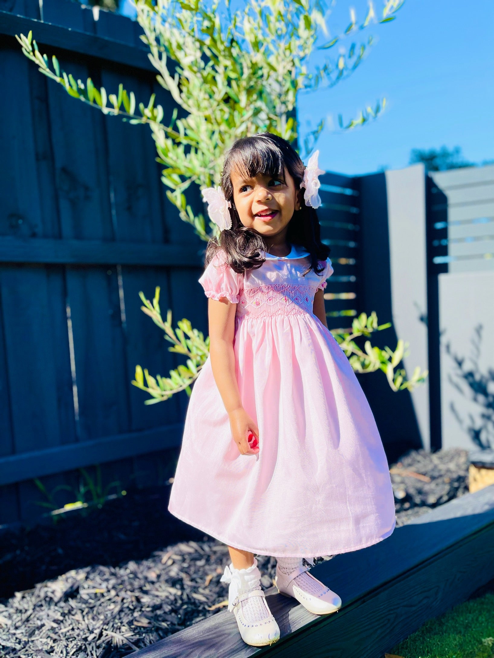 Girl toddler standing and looking side ways wearing the Hand-smocked Pink Sakura Dress. Light Pink Dress that has Pink and white handsmocking with dark pink rose bud embroidery. White collar with one pink rose bud embroidery on each collar. Dress is a light pink colour.
