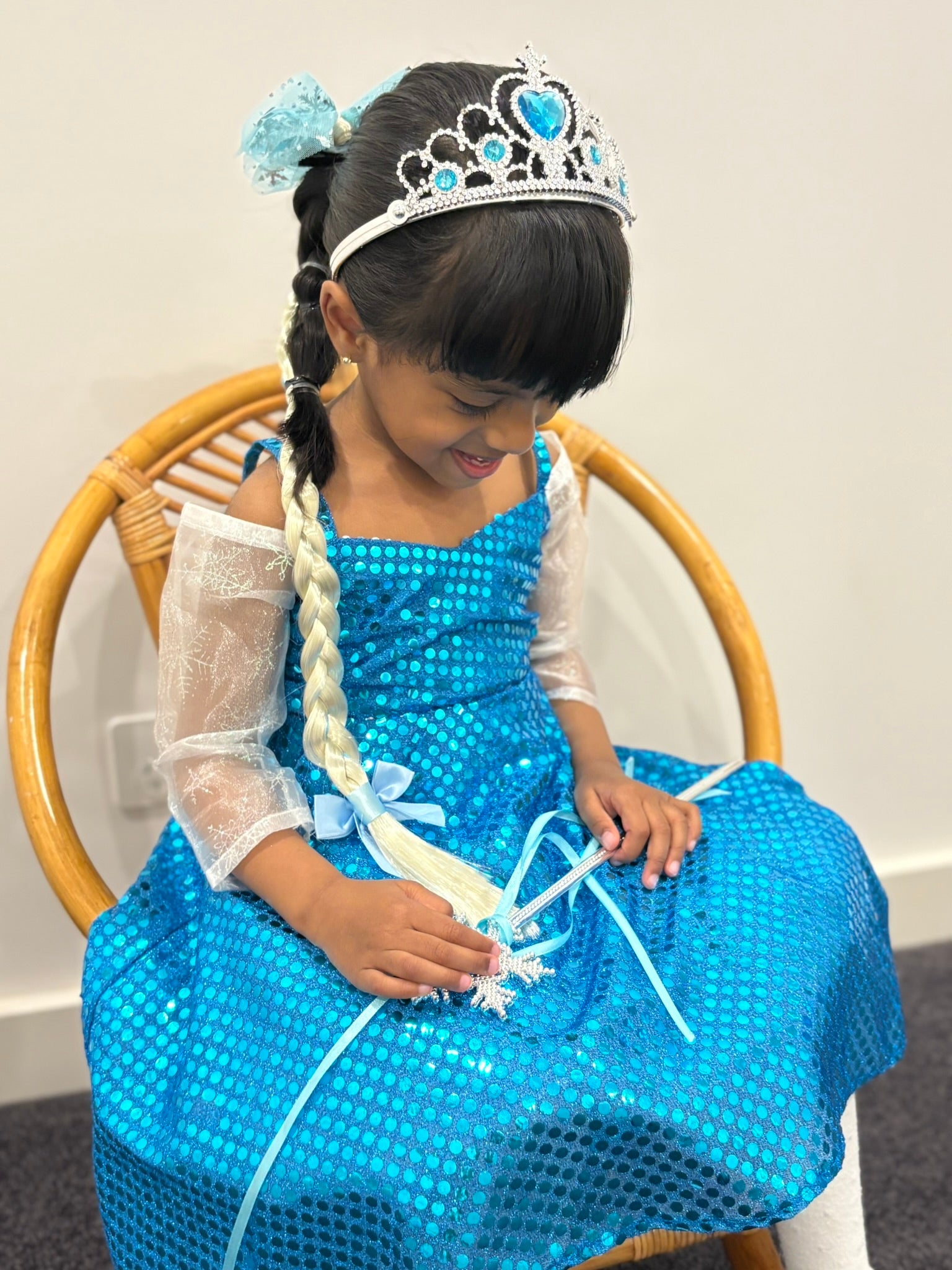 Child in a blue dress with a tiara sitting on a chair.