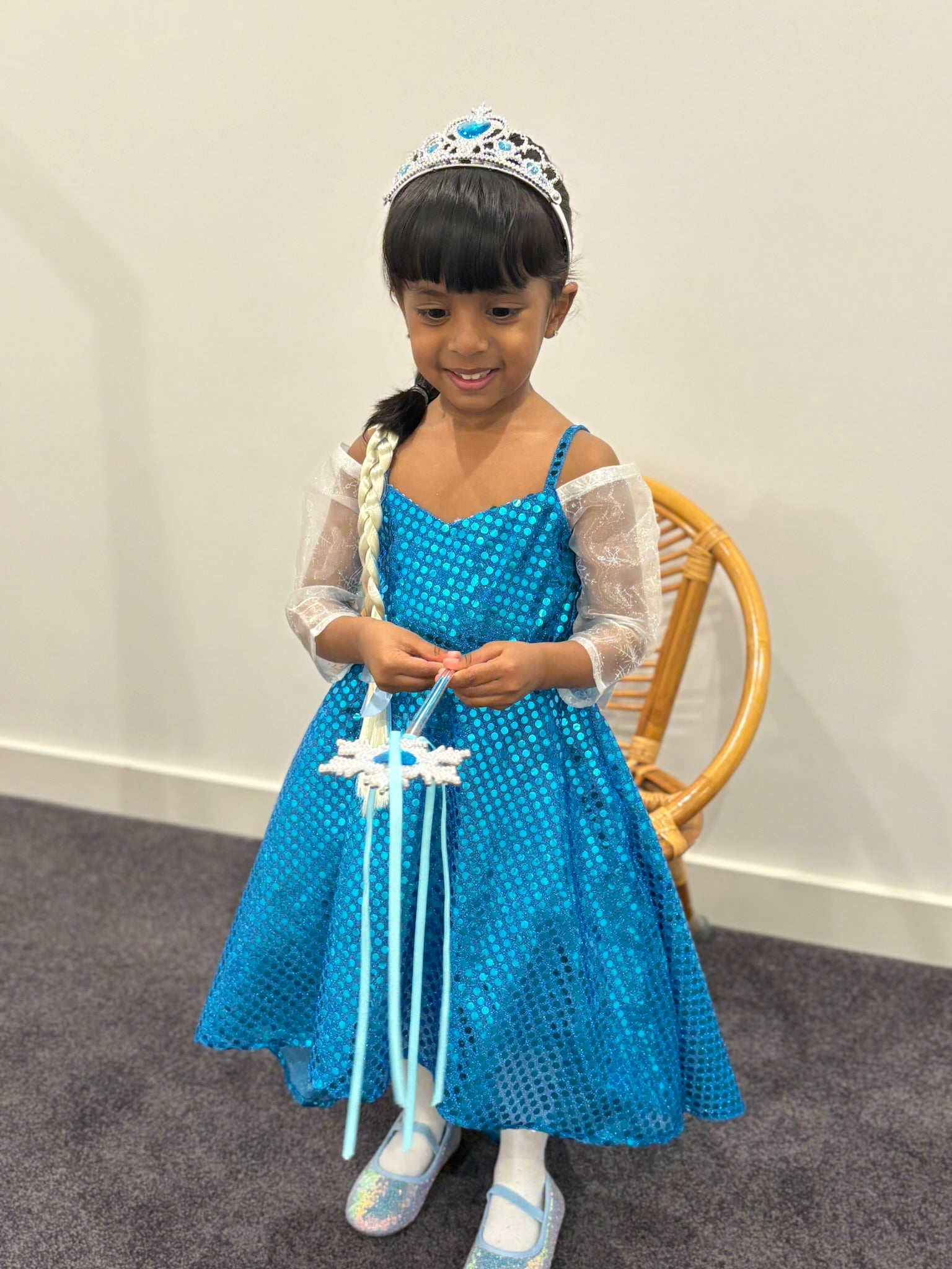 Young girl in a blue dress with a tiara and wand, sitting on a chair against a plain wall.