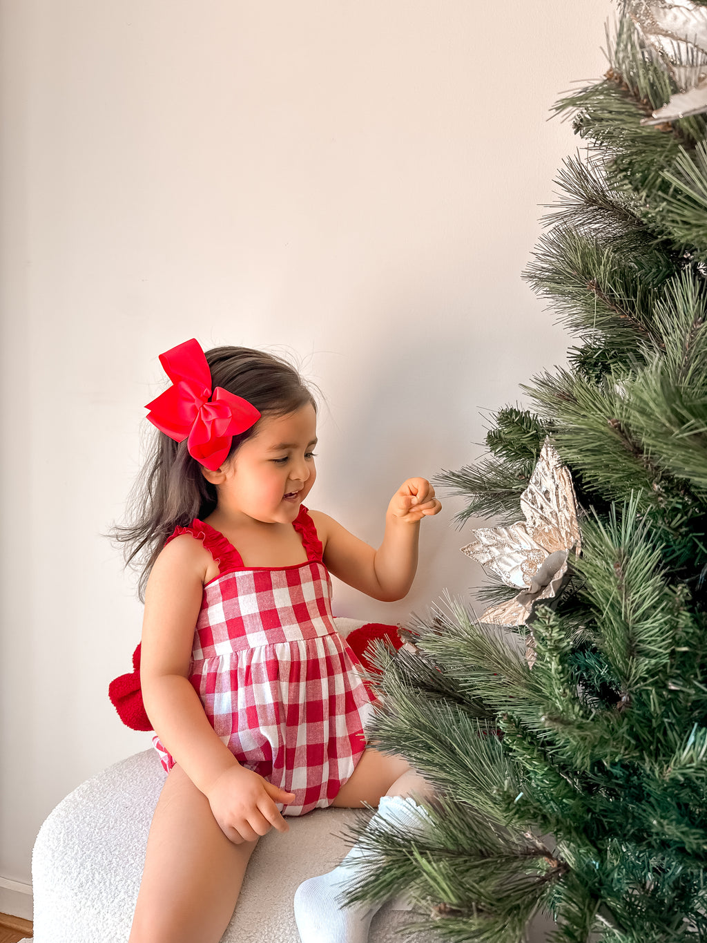 Child in a red and white checkered dress with a large bow sitting next to a Christmas tree.