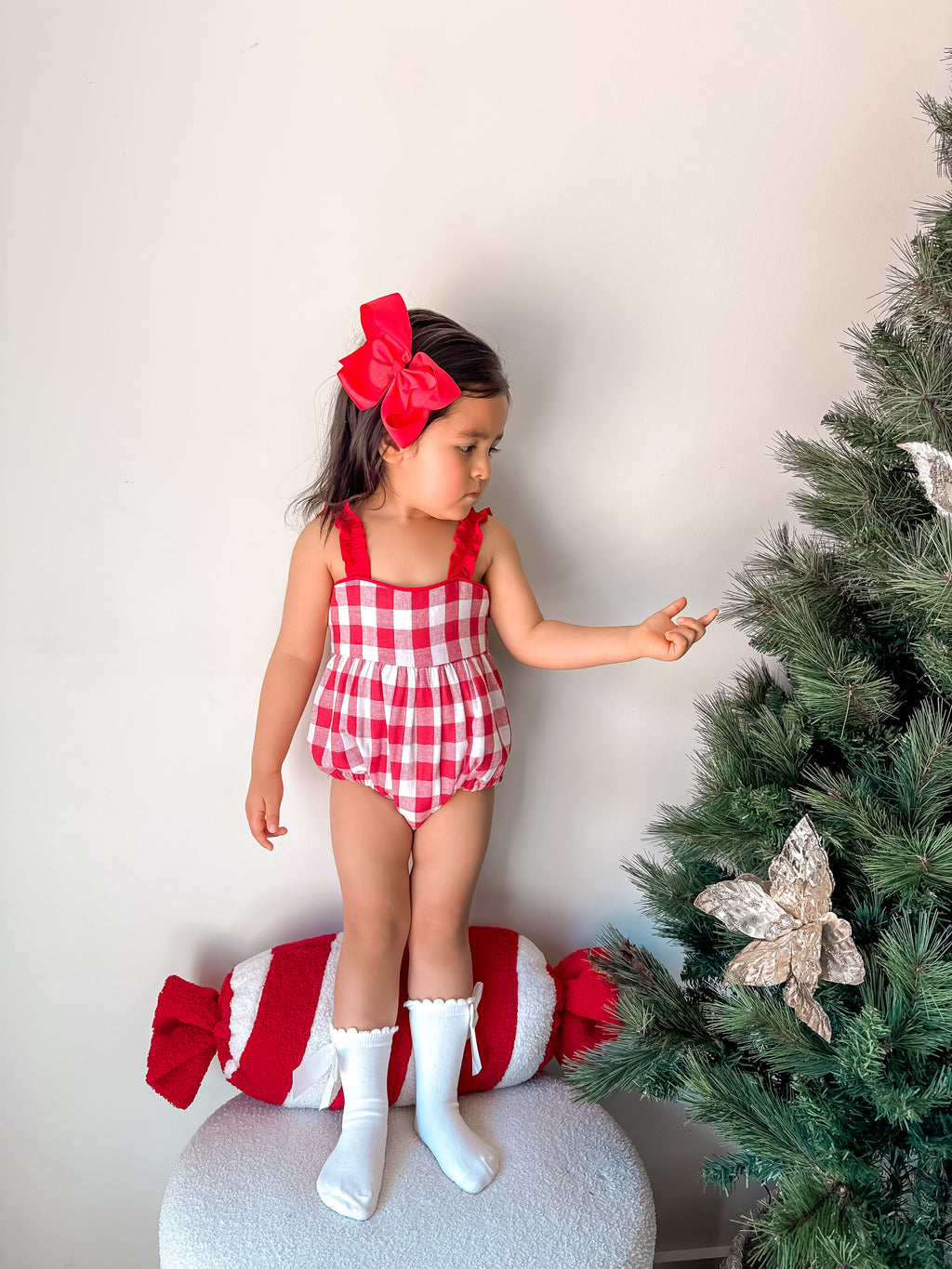 Child in red checkered outfit standing next to a decorated Christmas tree.