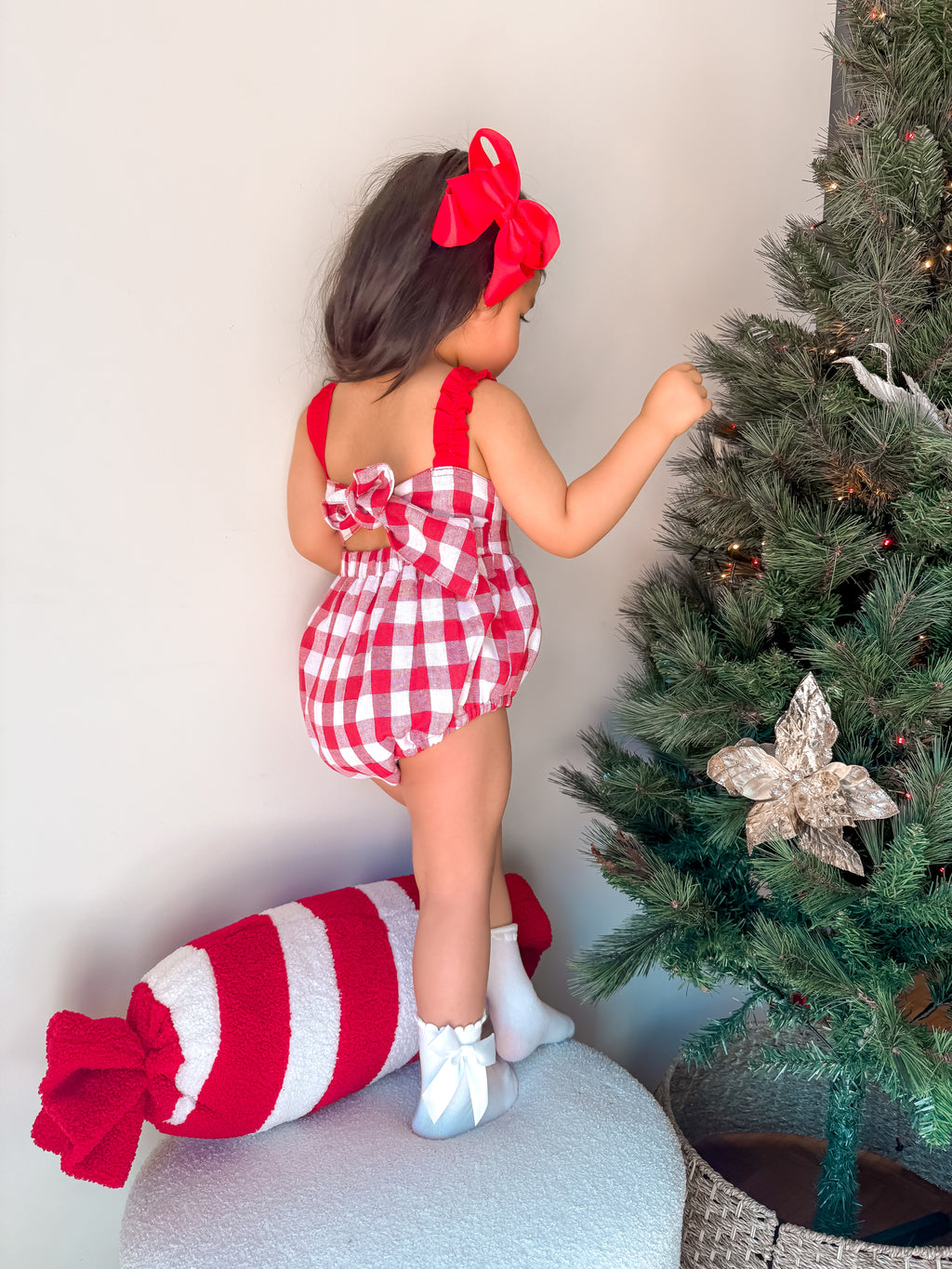 Child in a red and white checkered outfit standing next to a decorated Christmas tree.
