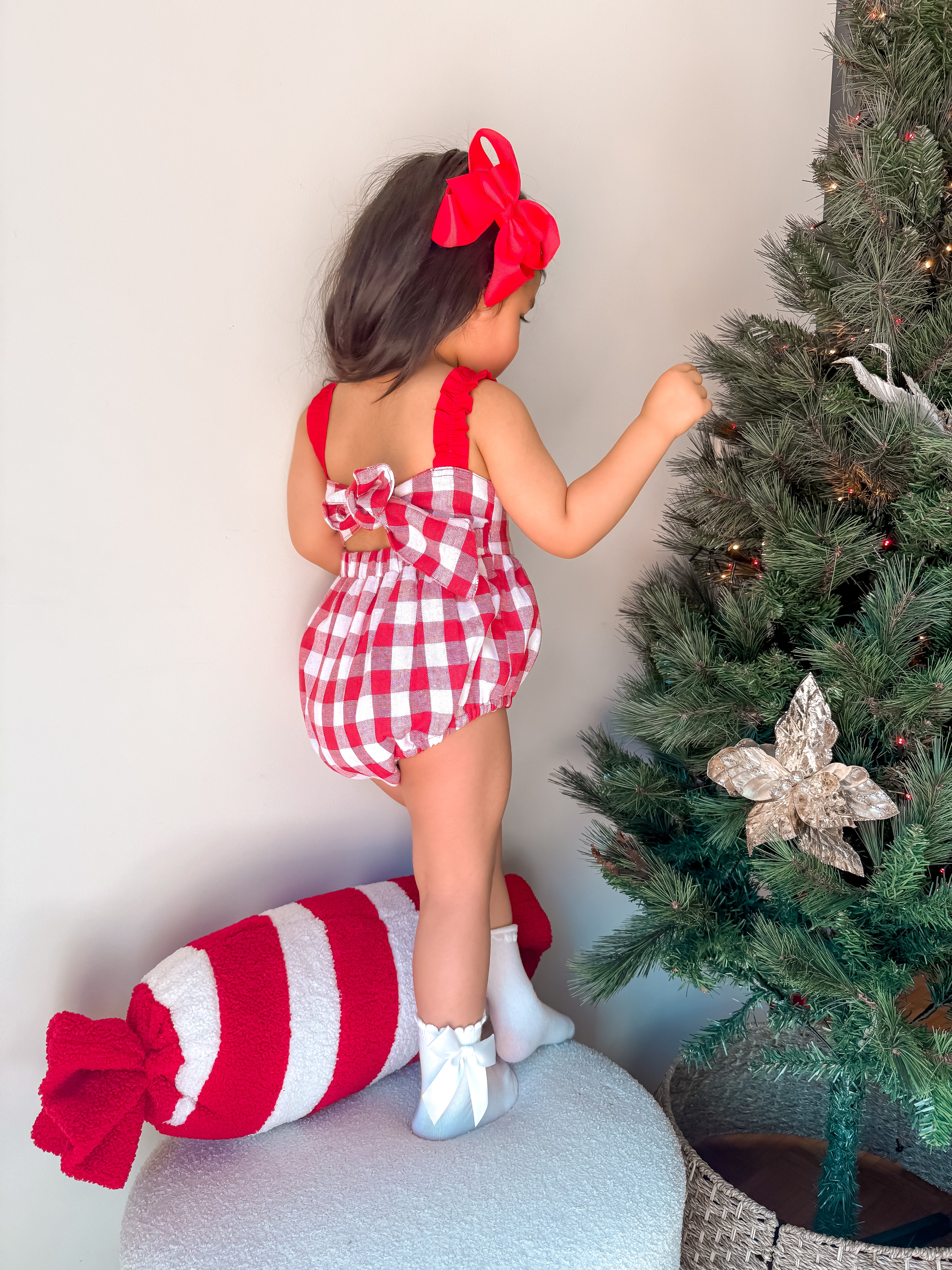 Child in a red and white checkered outfit standing next to a decorated Christmas tree.