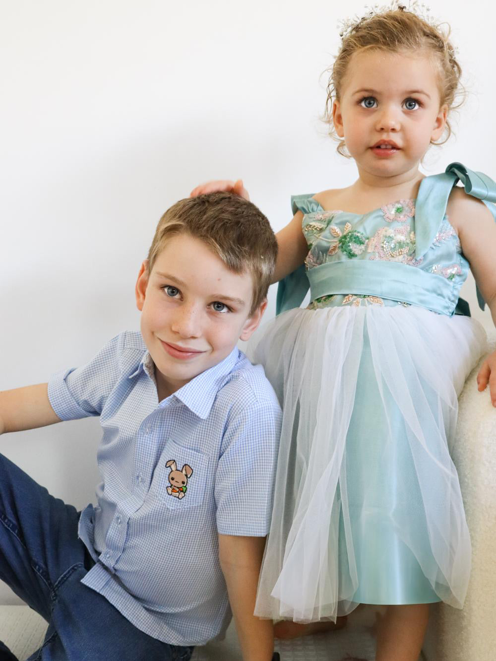 Two children, a boy and a girl, sitting together on a white background. boy in a white and blue gingham shirt made of cotton with a cute embroidered bunny on the side pocket. short sleeve