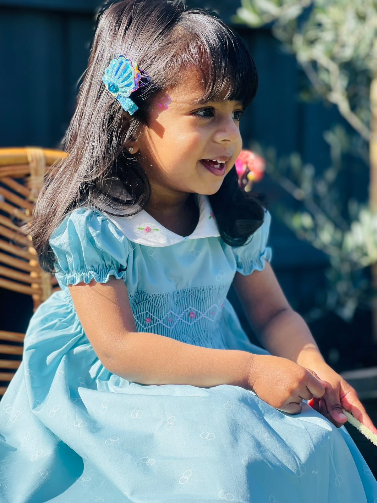 Girl sitting-Bluebell hand-smocked dress in blue with white collar and pink rose bud embroidery and blue white and pink handsmocking