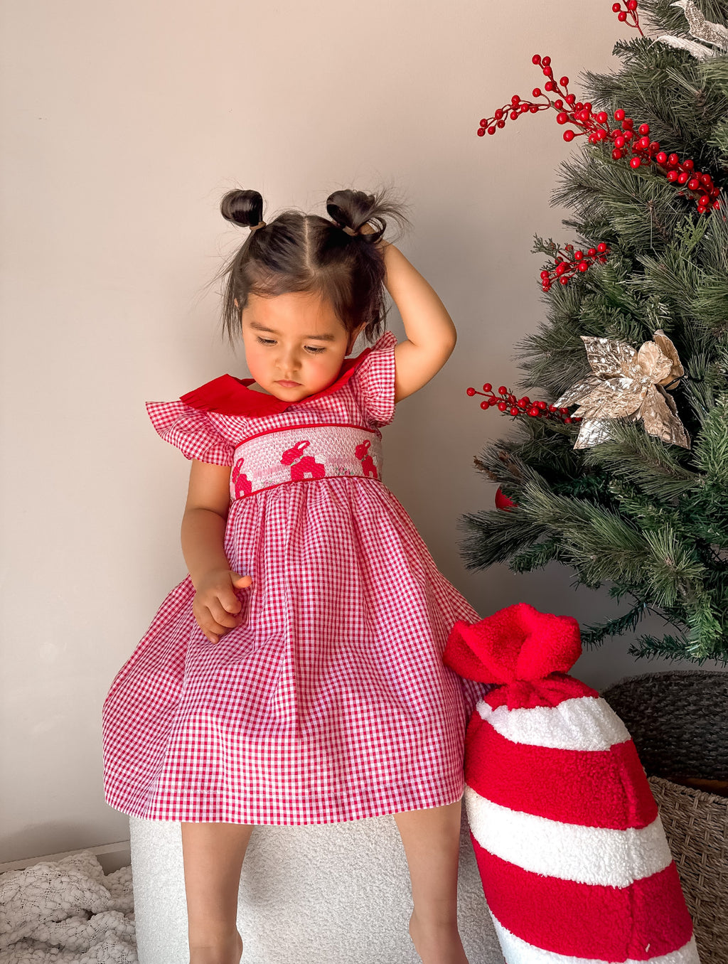 Child in a red and white checkered dress standing next to a decorated Christmas tree.