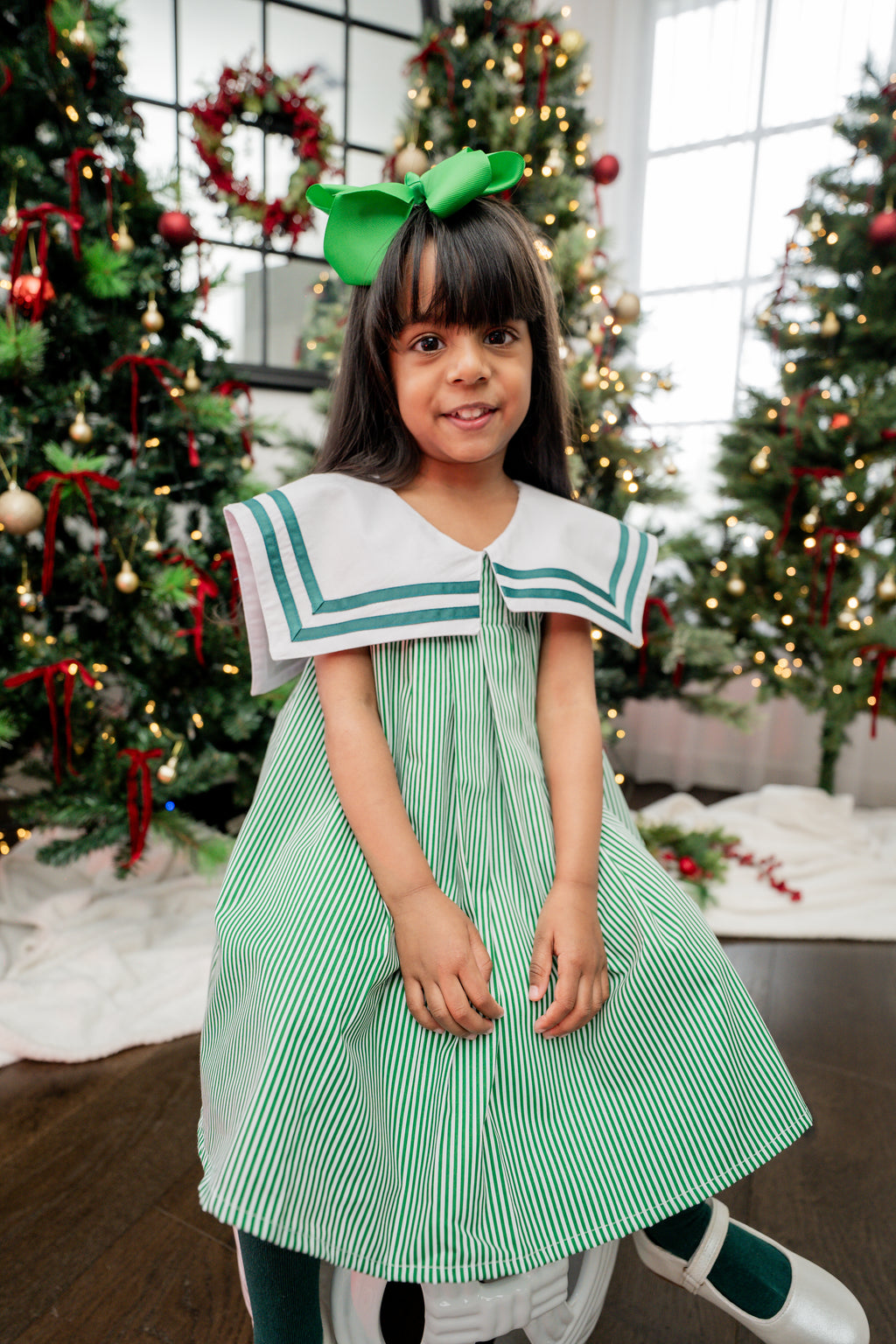Young girl in a green and white dress with a large bow in front of Christmas trees.