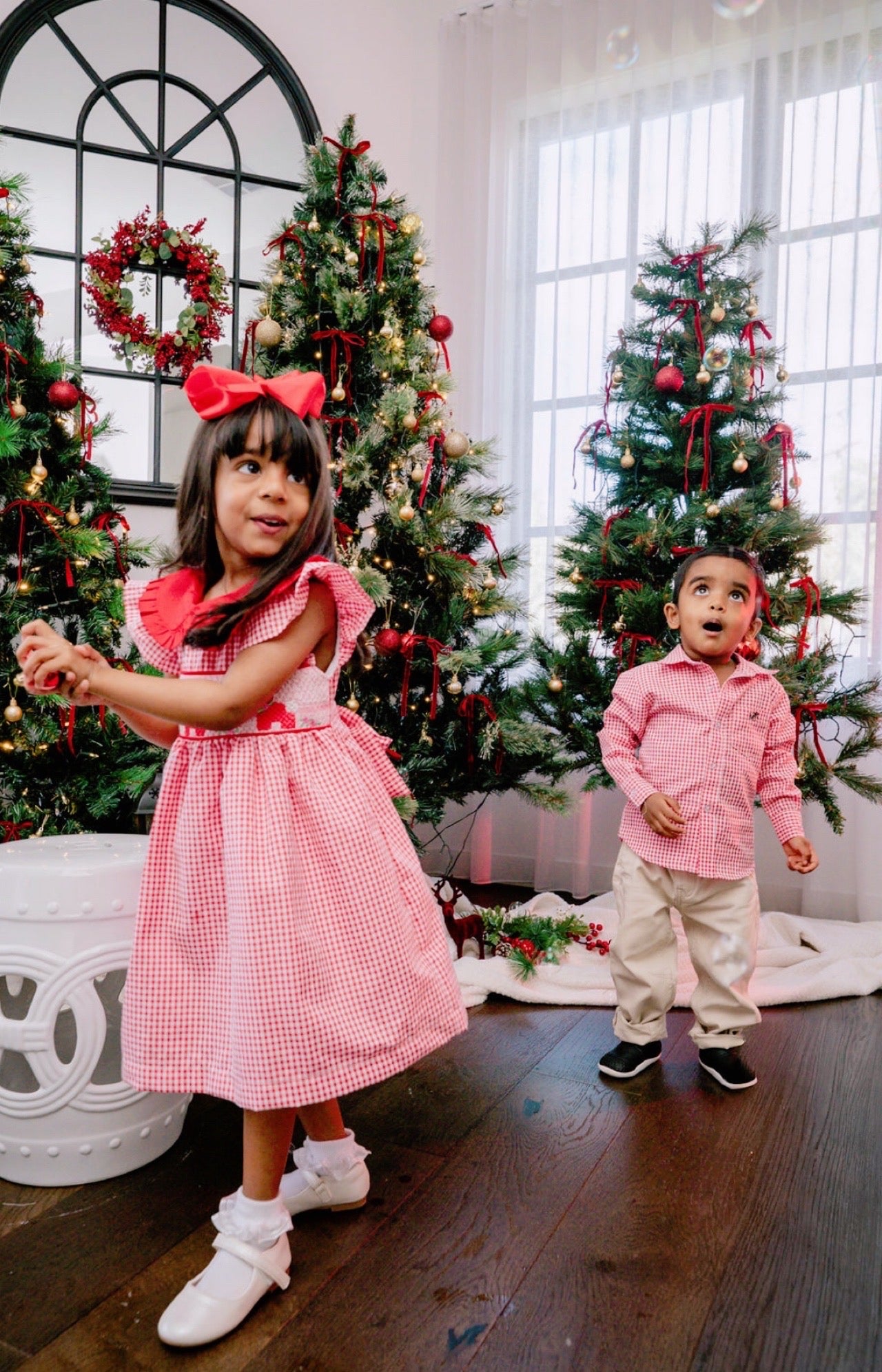 Two children in festive attire standing in a room decorated with Christmas trees.