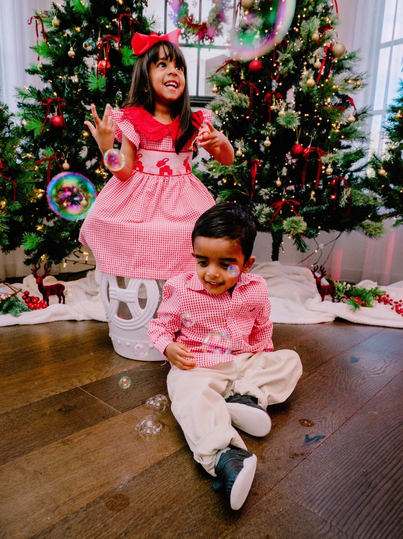 Two children in matching outfits sitting on the floor with Christmas trees in the background.