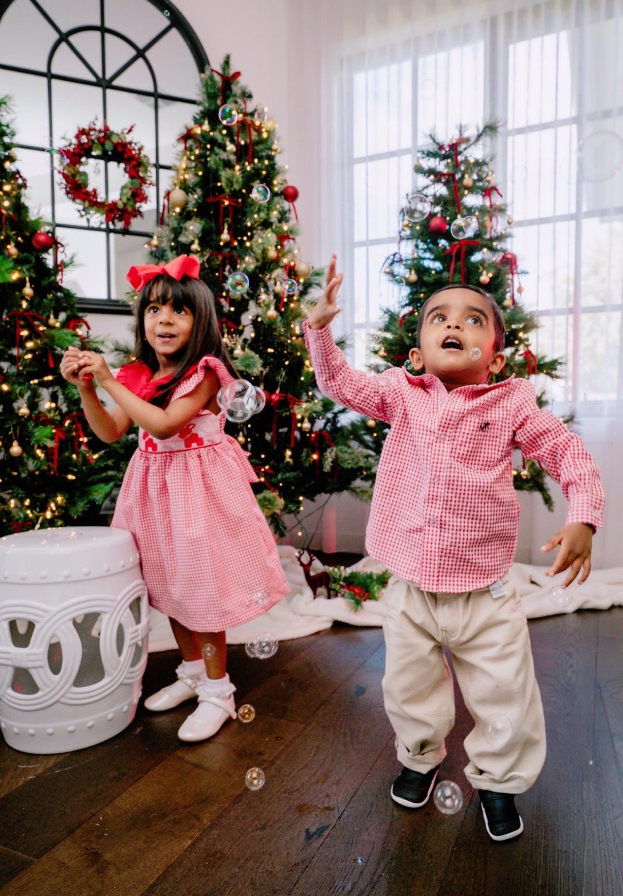 Two children in festive attire standing in a room decorated with Christmas trees.