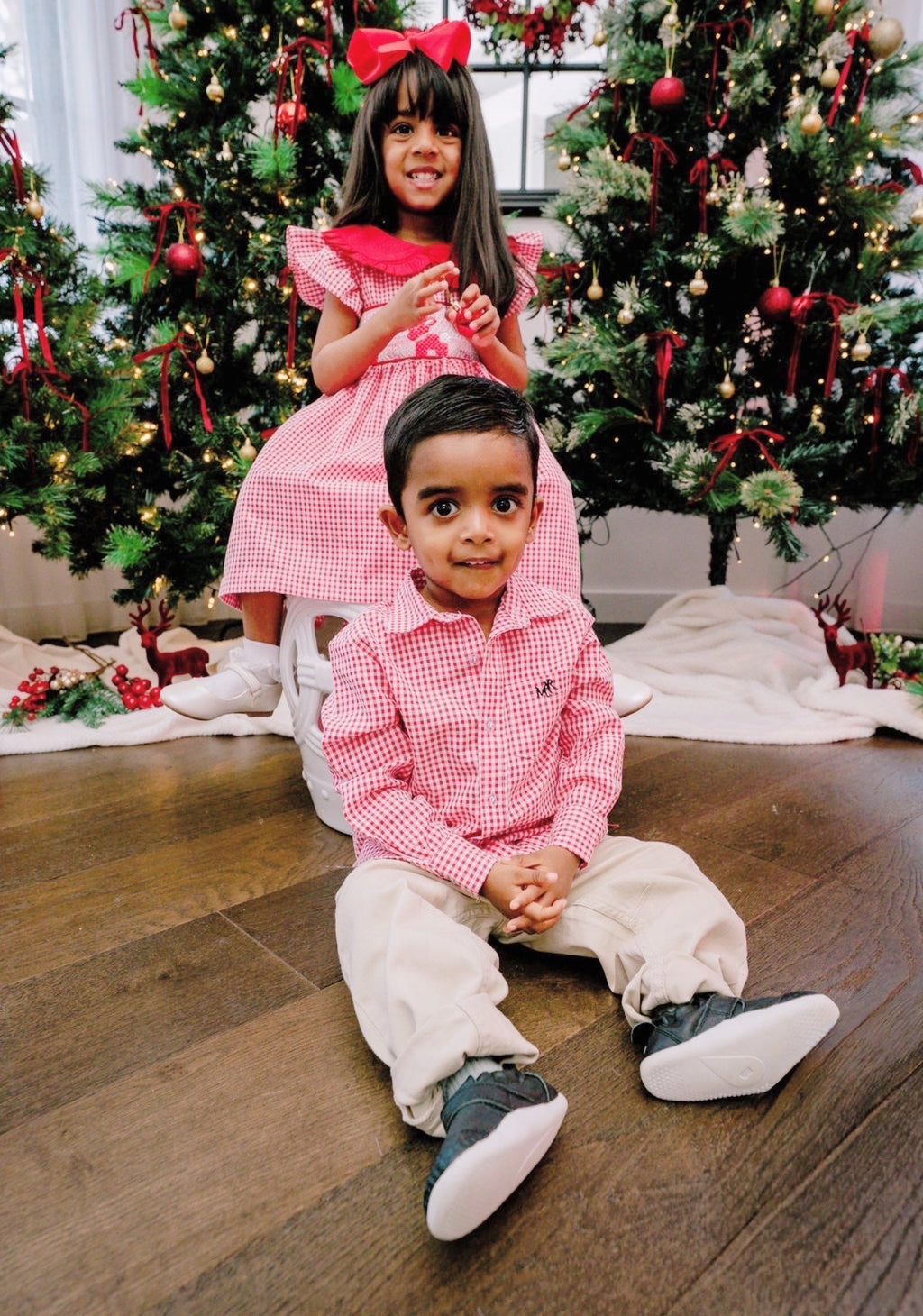 Two children in matching outfits sitting on a wooden floor with Christmas trees in the background.