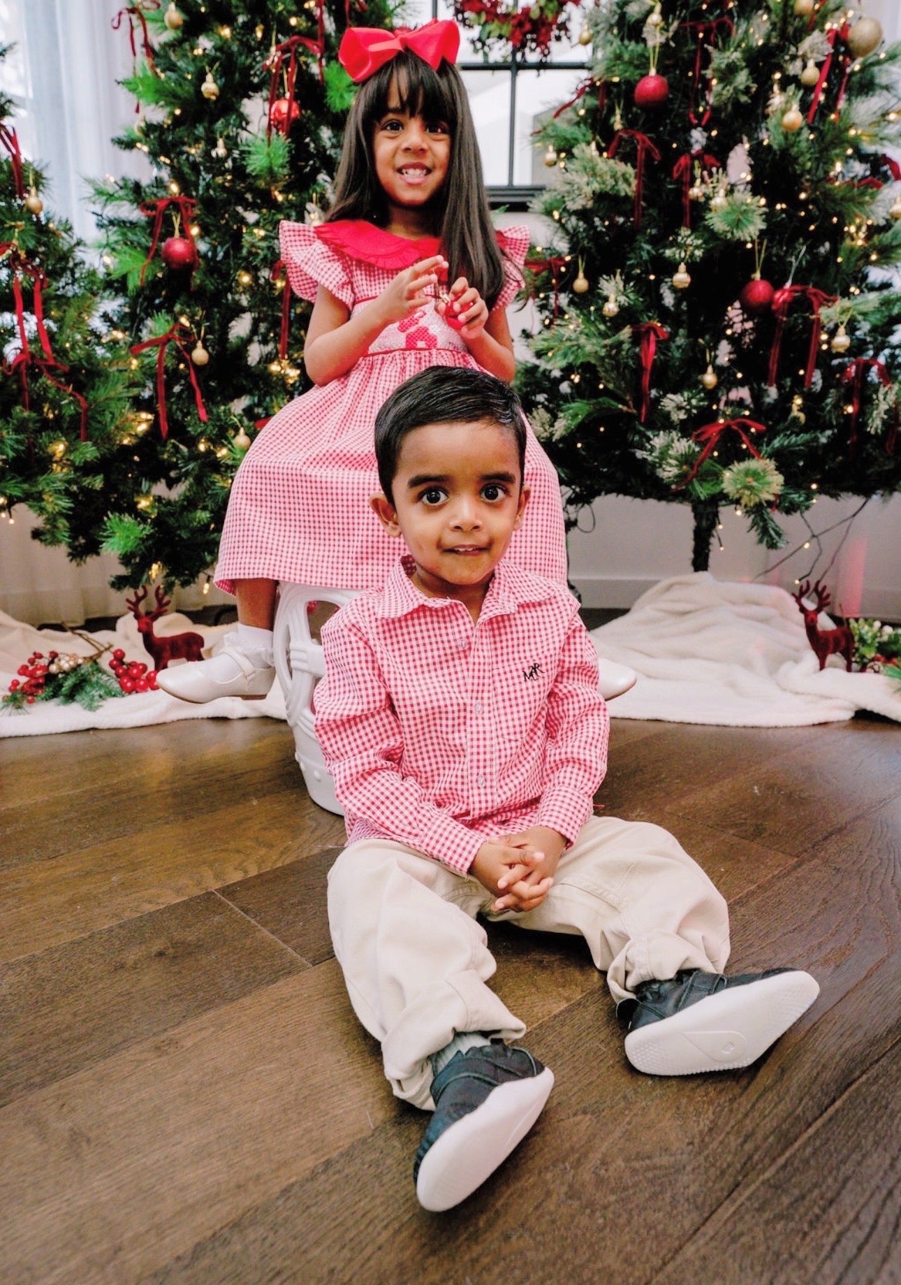 Two children in matching outfits sitting on a wooden floor with Christmas trees in the background.
