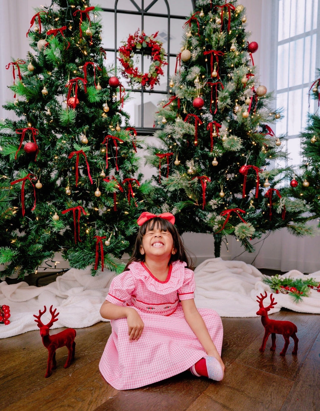 Child in a pink dress sitting in front of decorated Christmas trees with red ribbons.