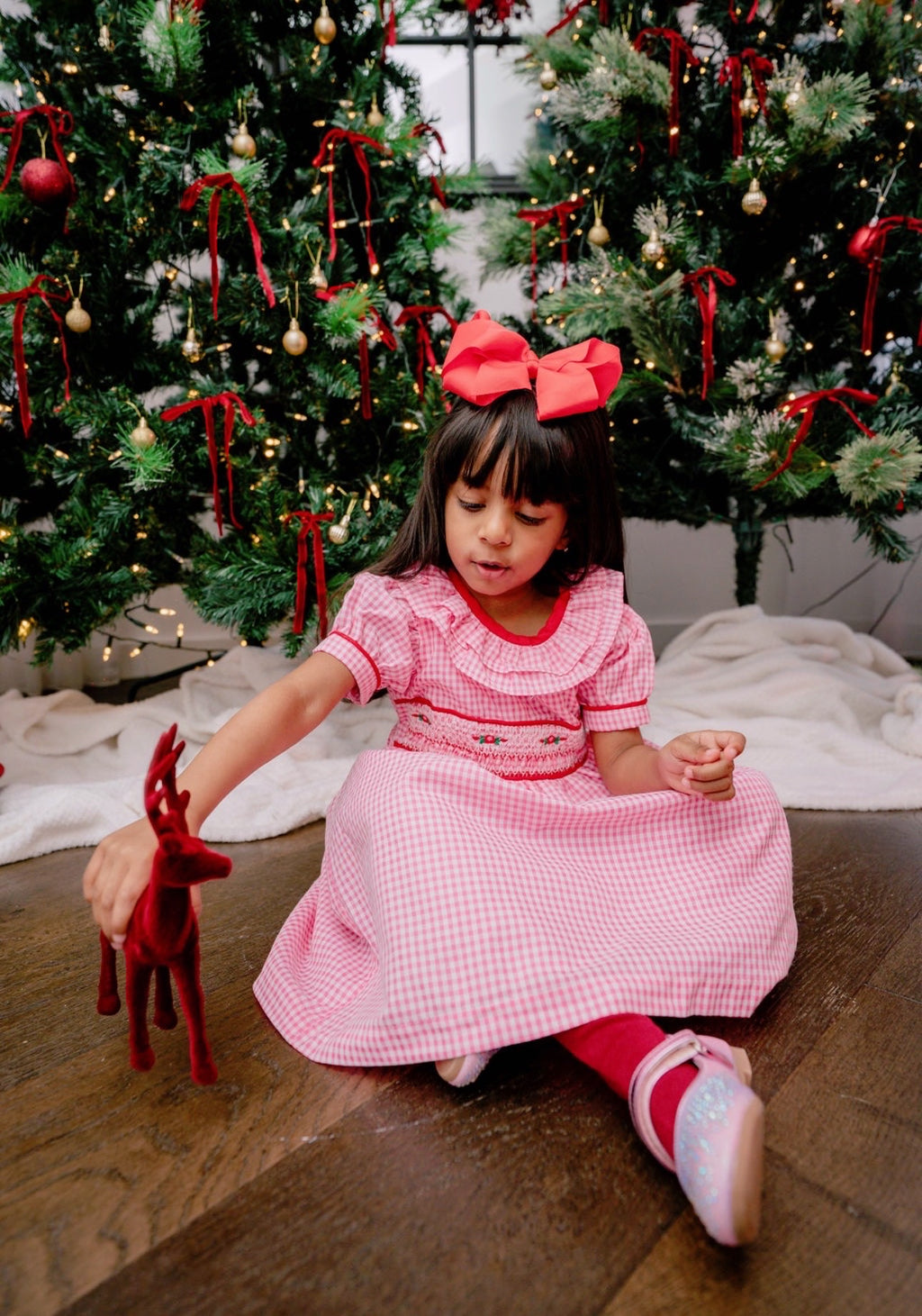 Young girl in a pink dress sitting on the floor with Christmas trees in the background