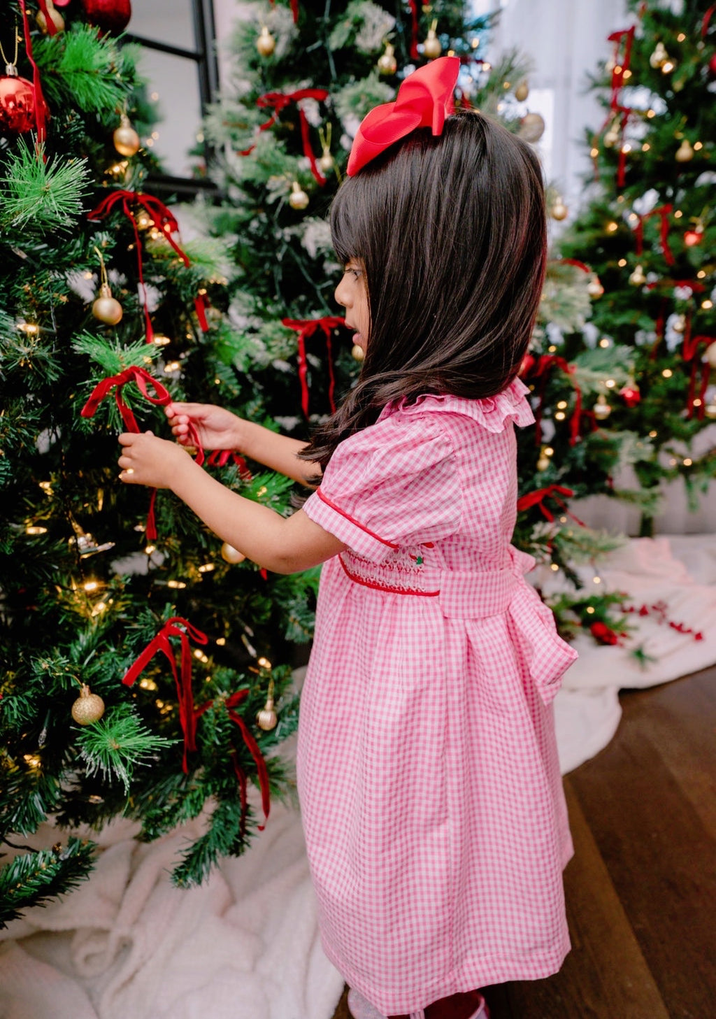 Child in a pink dress decorating a Christmas tree with red ribbons.