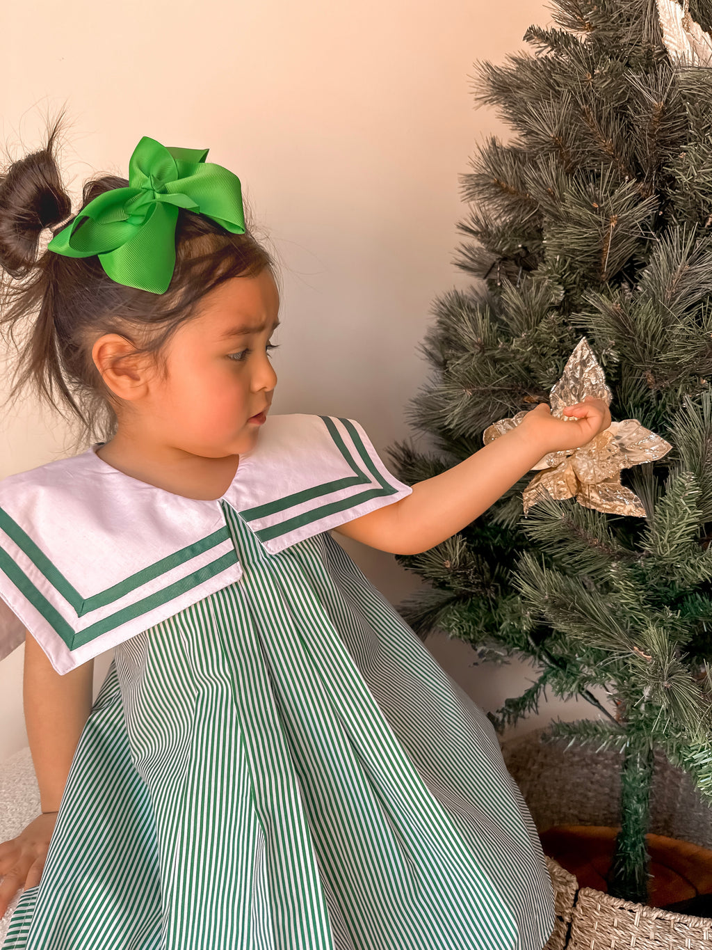 Child in a green and white dress with a large bow, decorating a Christmas tree.