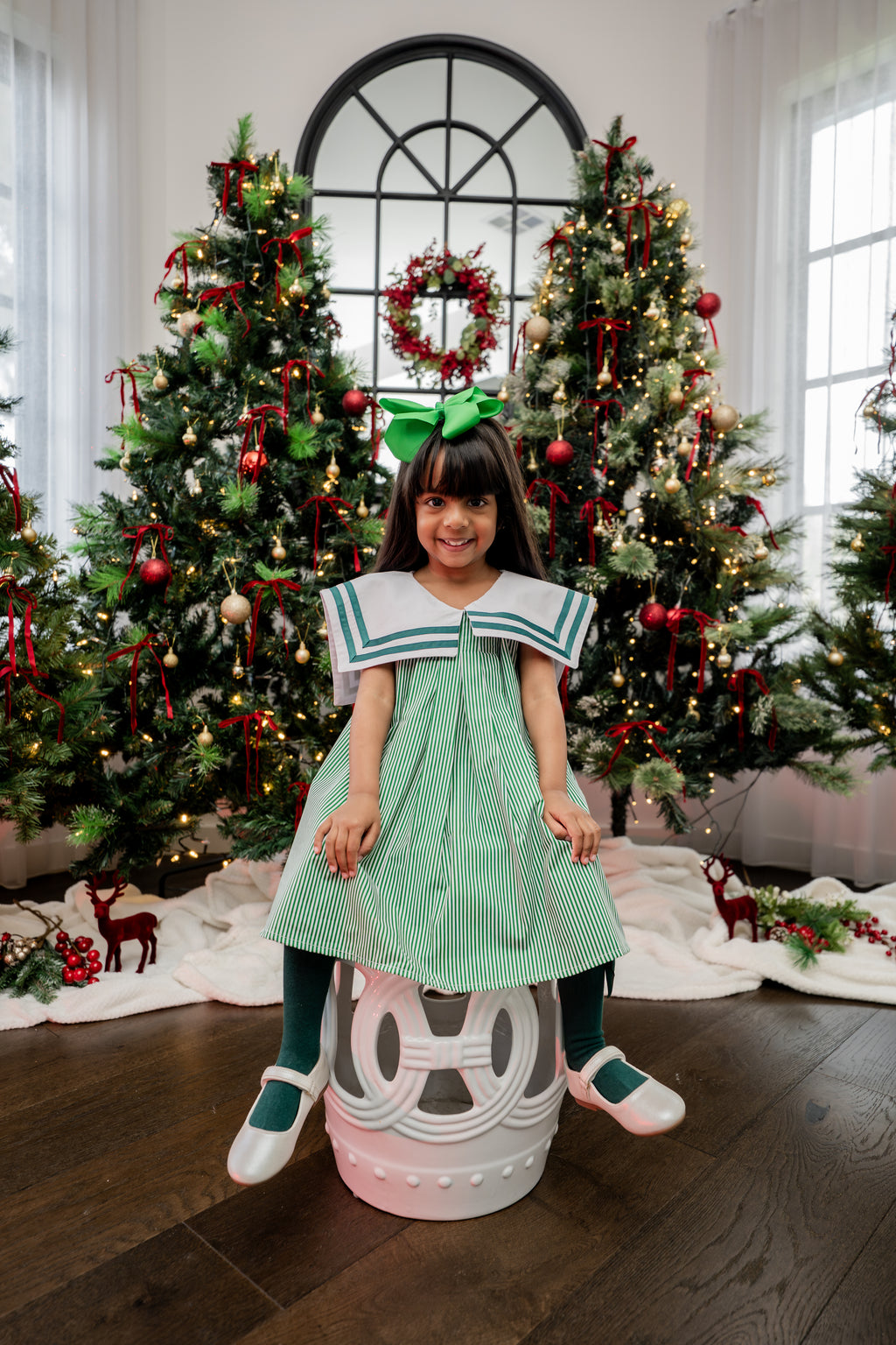 Child in a green dress standing in front of Christmas trees with decorative lights and wreaths.